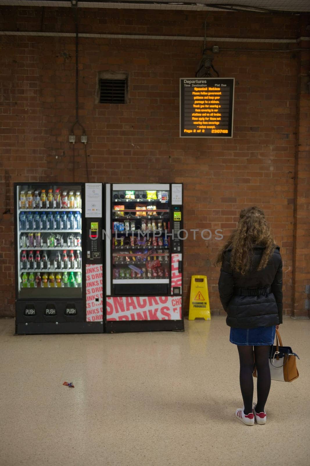Doncaster, United Kingdom, 22nd May, 2021: One young woman waits patiently near vending machines, holding hand bag, looking at train departure board