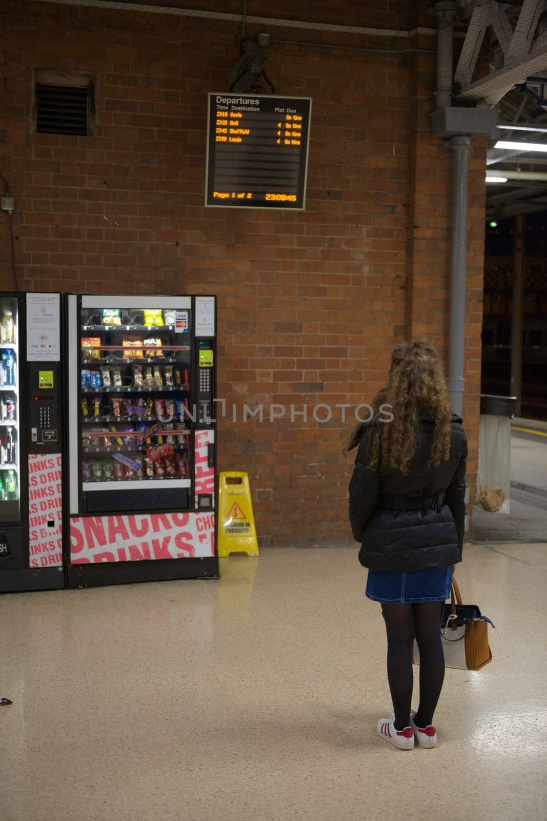 Doncaster, United Kingdom, 22nd May, 2021: Vertical image of a winter clothed young woman reading doncaster train station departure board. Late night