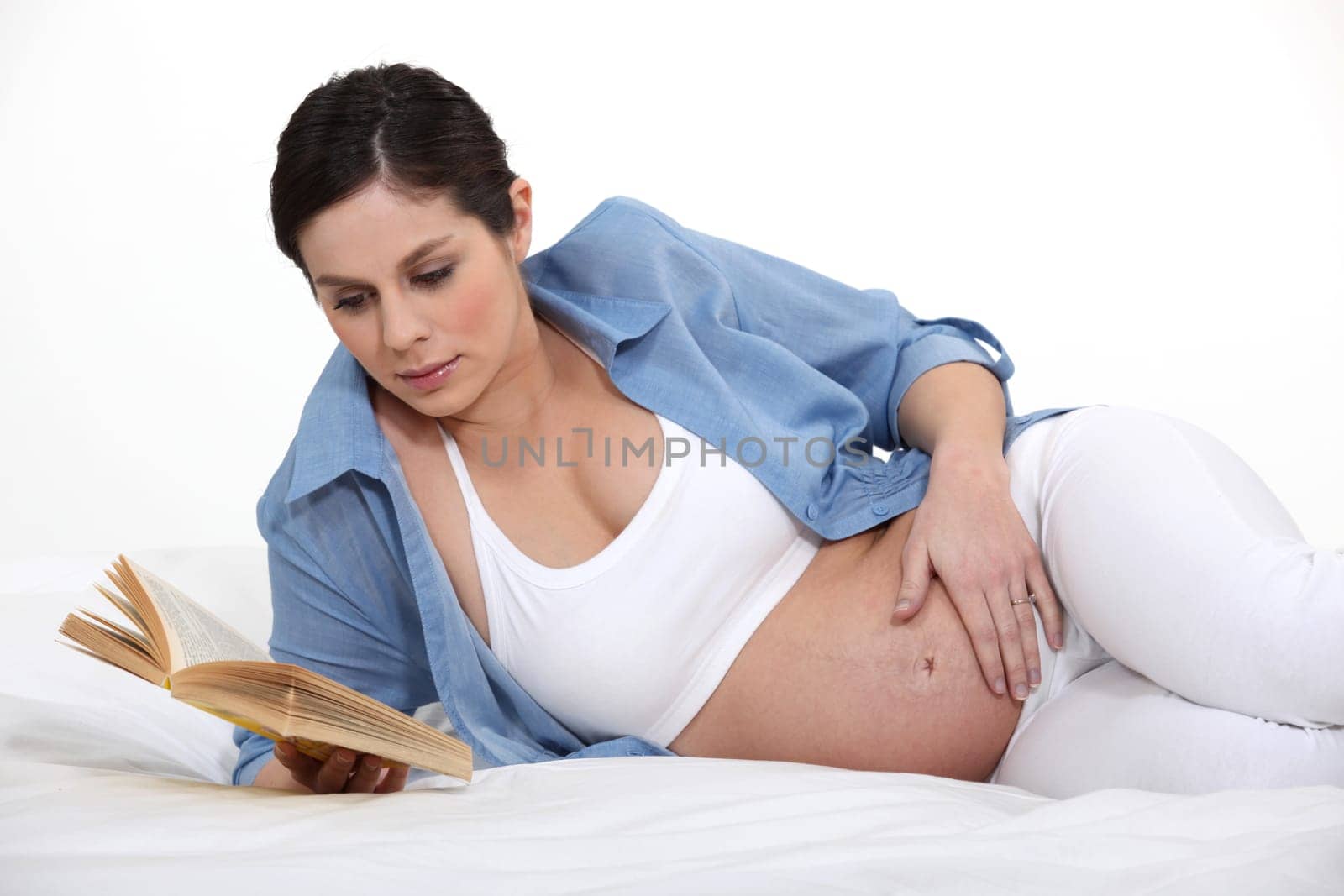 A pregnant woman reading a book on her bed.