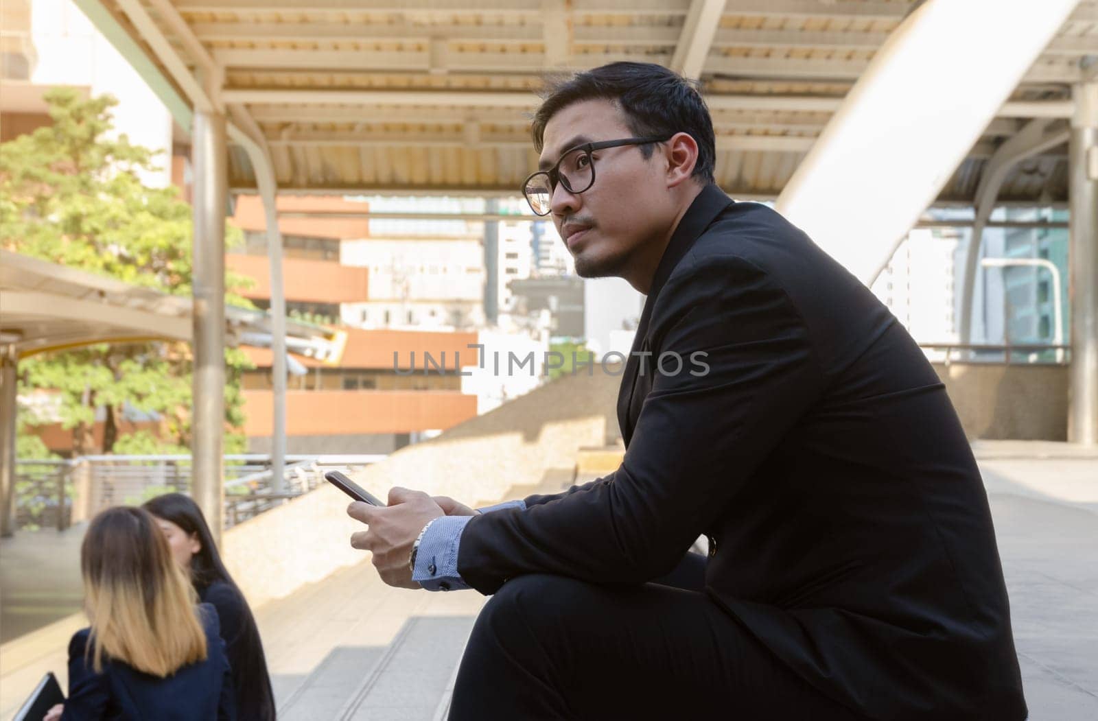 Thoughtful businessman holding smartphone and looking away . Stress man wearing glasses and black suit with serious expression sitting on steps outdoors in the city. Technology and business concept