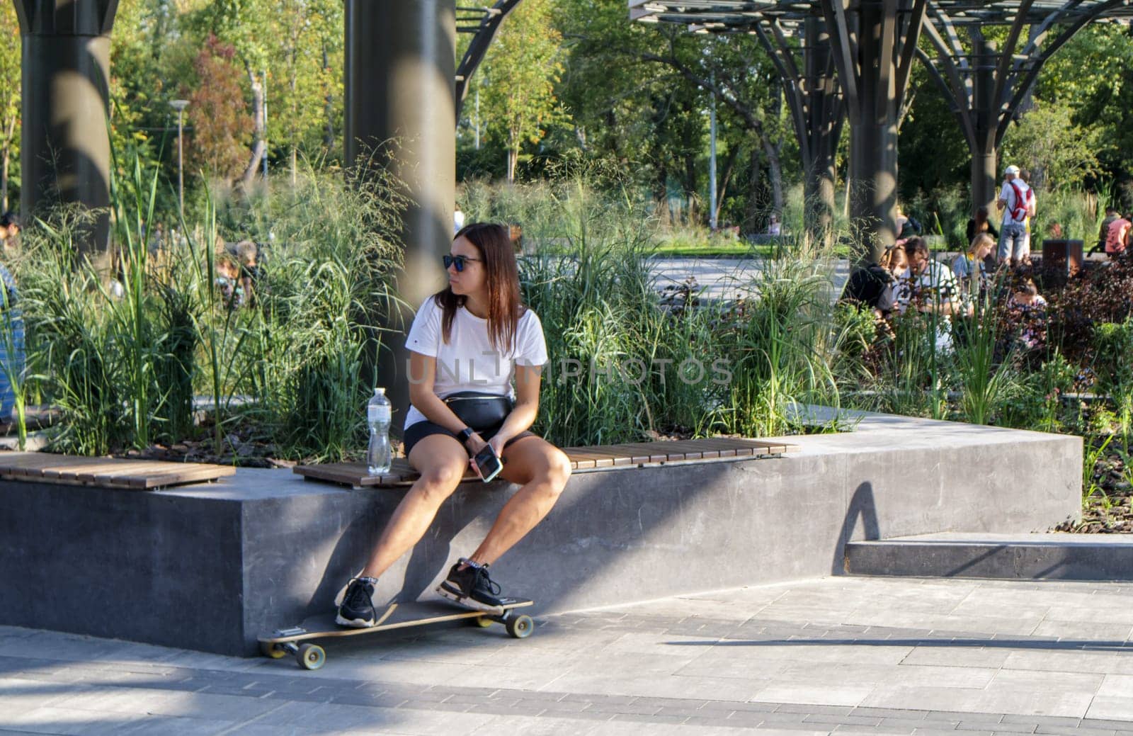 Young caucasian girl with a skateboard, sunglasses outdoors sits on the parapet and relaxes after skating. summer outdoor activities. Ukraine, Kiev - September 13, 2021