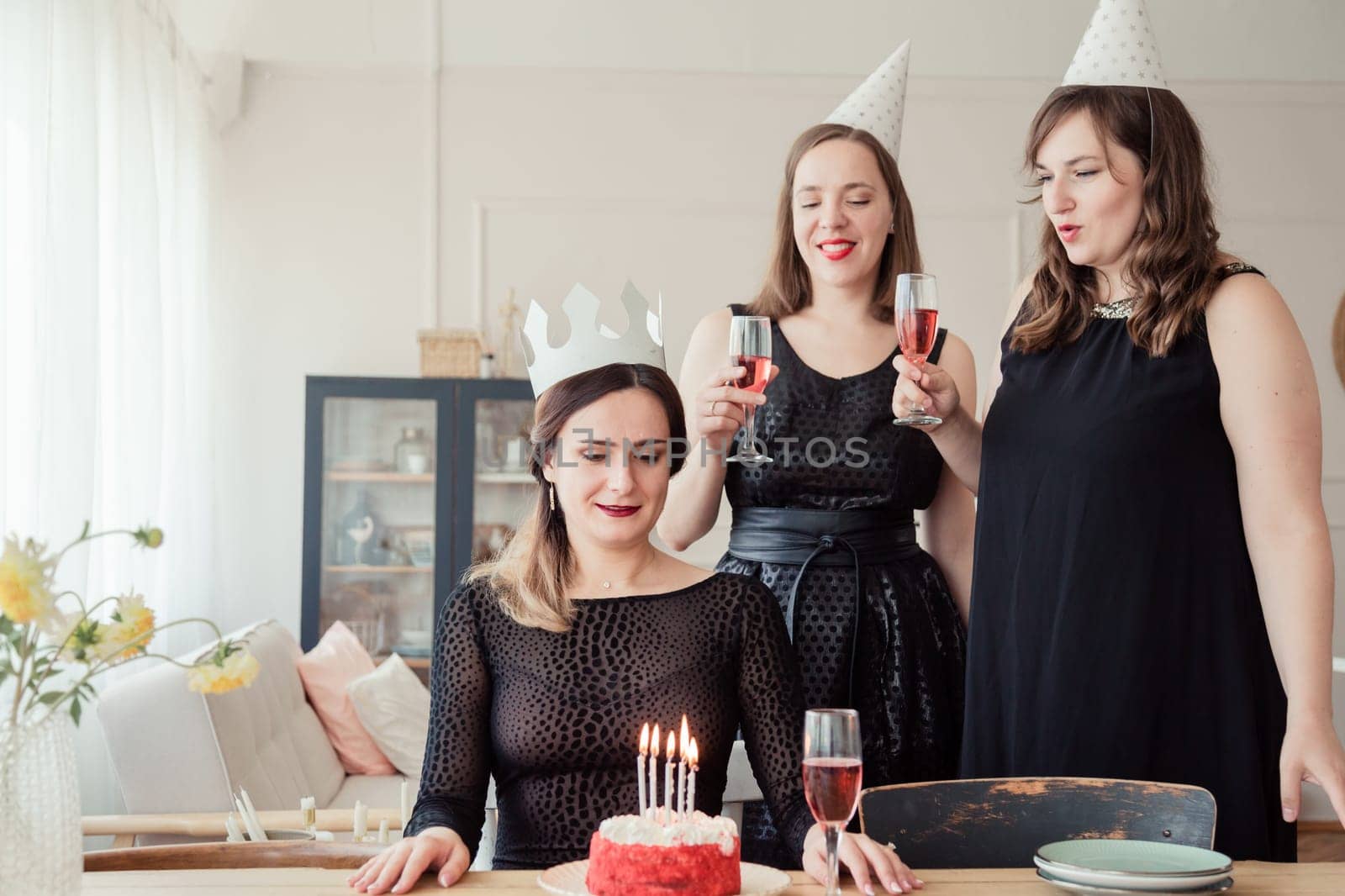 Three women friends celebrating 30s birthday together gathering at table at home with wine and birthday cake with candles. Women in party hat and birthday girl in crown blows out candles