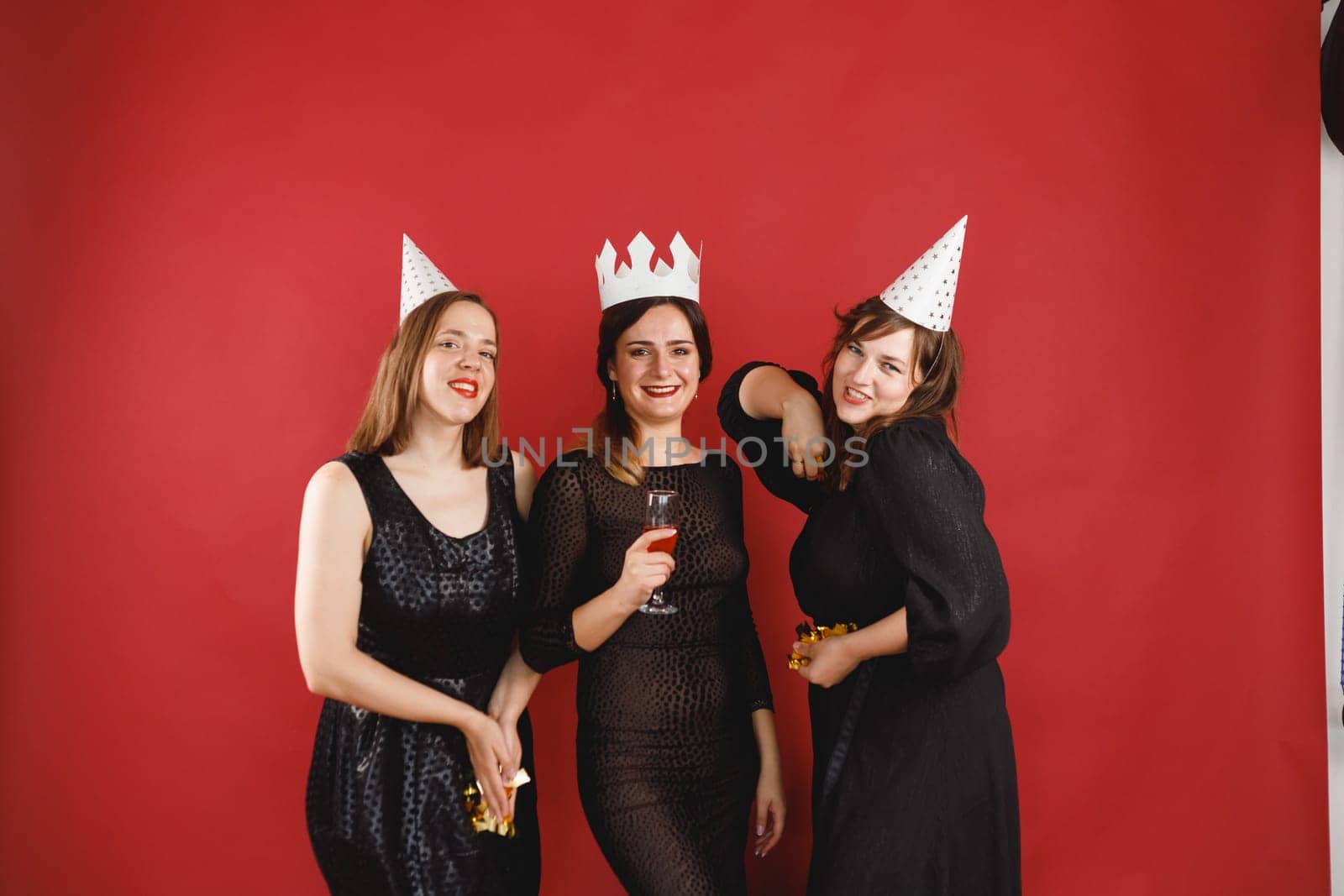 Three delighted women in black dress and party hats over red background with golden confetti. Female party