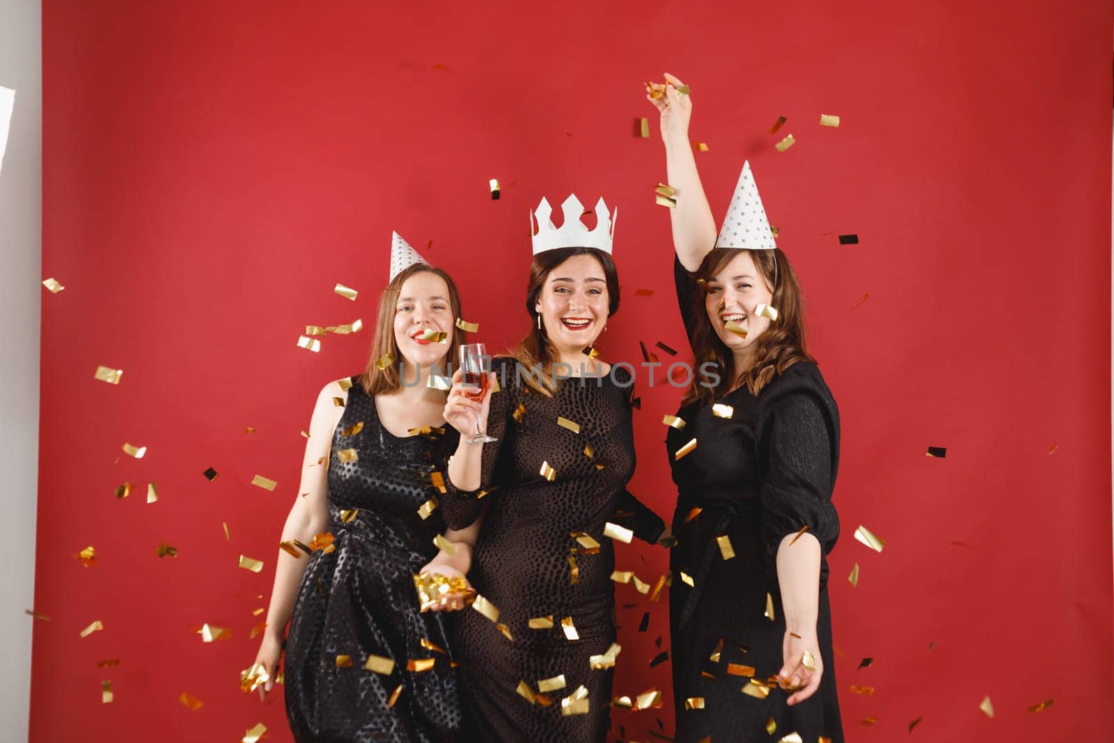 Three delighted women in black dress and party hats over red background with golden confetti. Female party