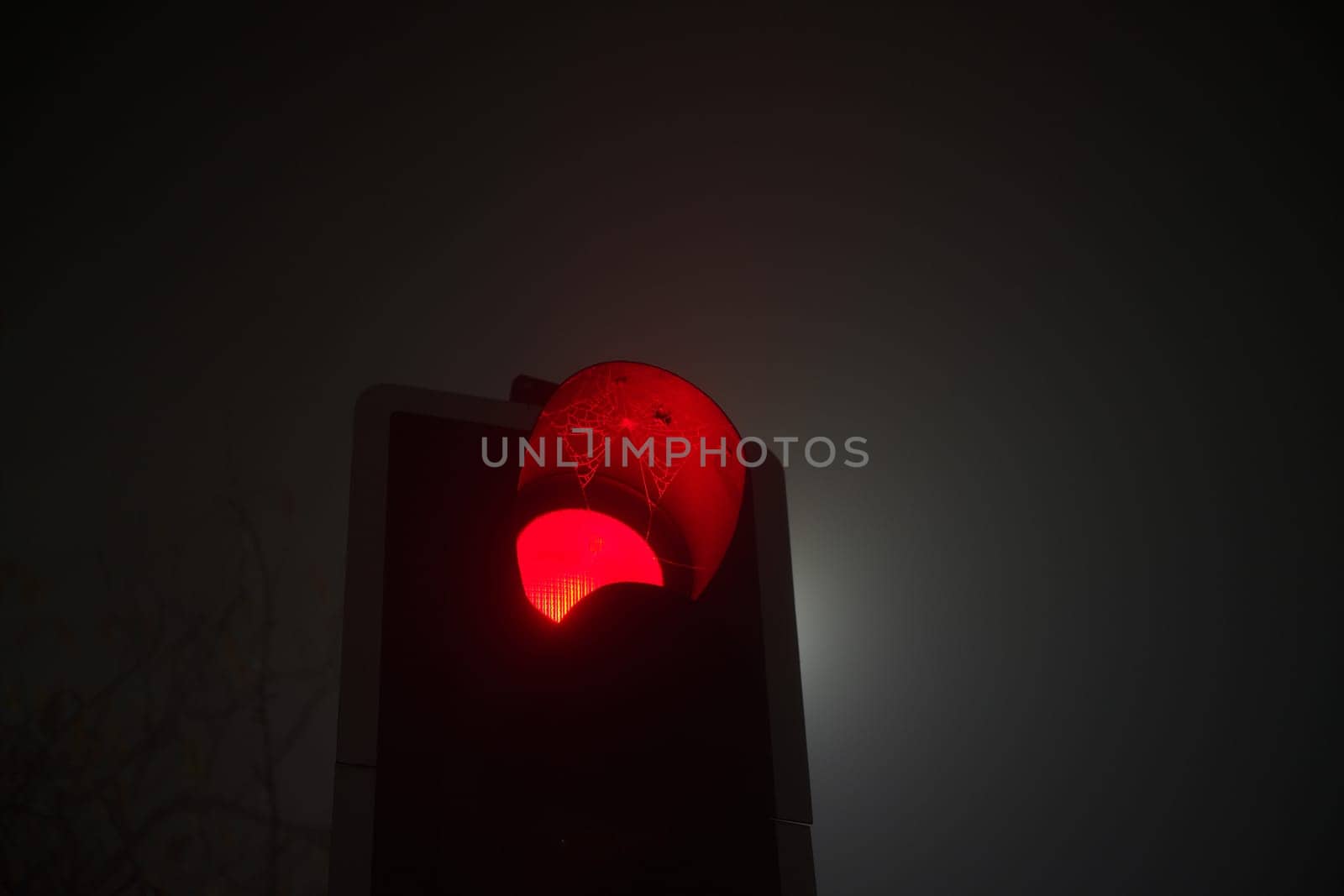 A british traffic light shines red, indicating stop, during foggy conditions at night. Also, a cobweb hangs over the red light of the traffic signal