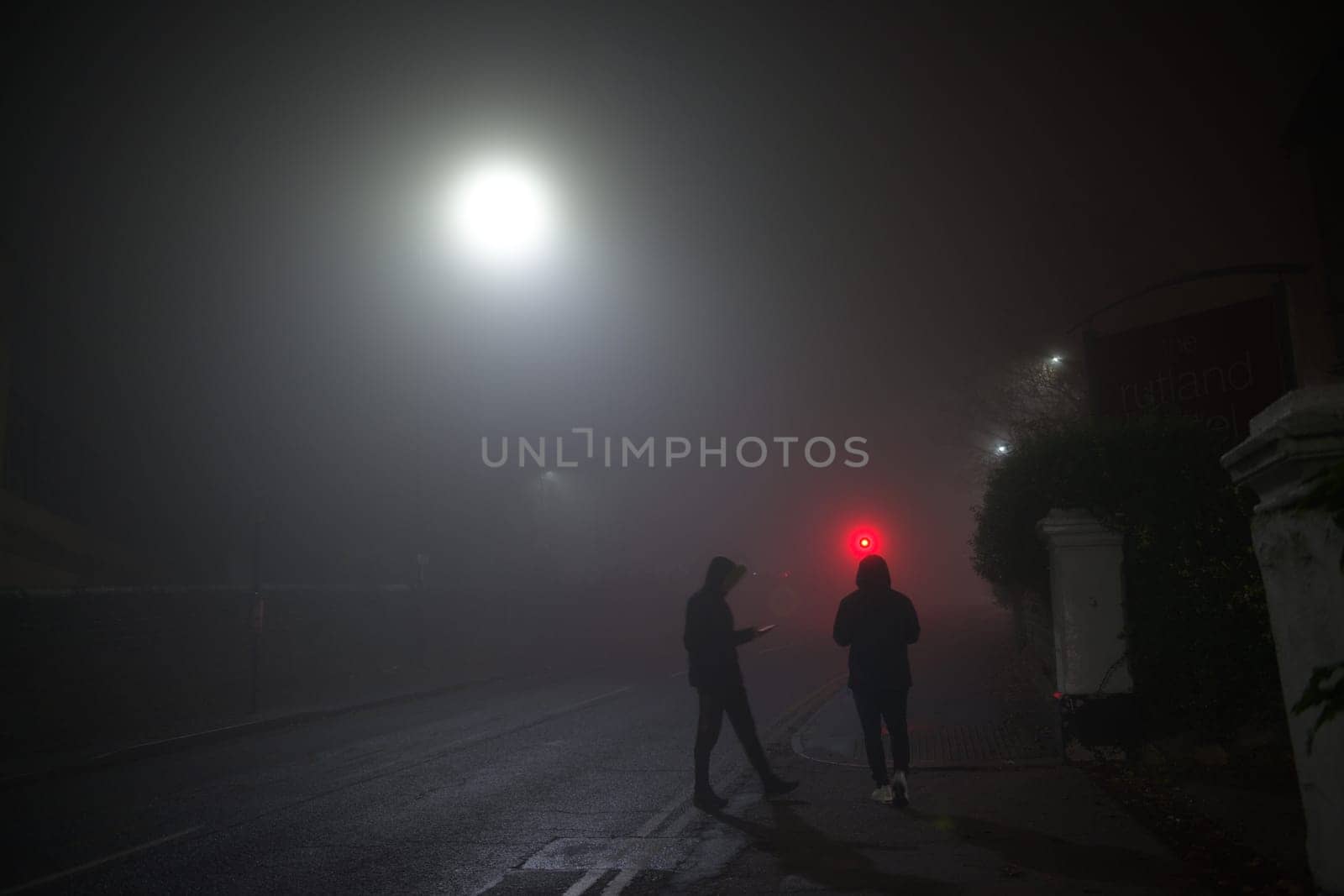 Hooded youths loiter on a street corner using mobile smartphones. Dark and foggy conditions with urban setting. Both wear winter clothes with trainers