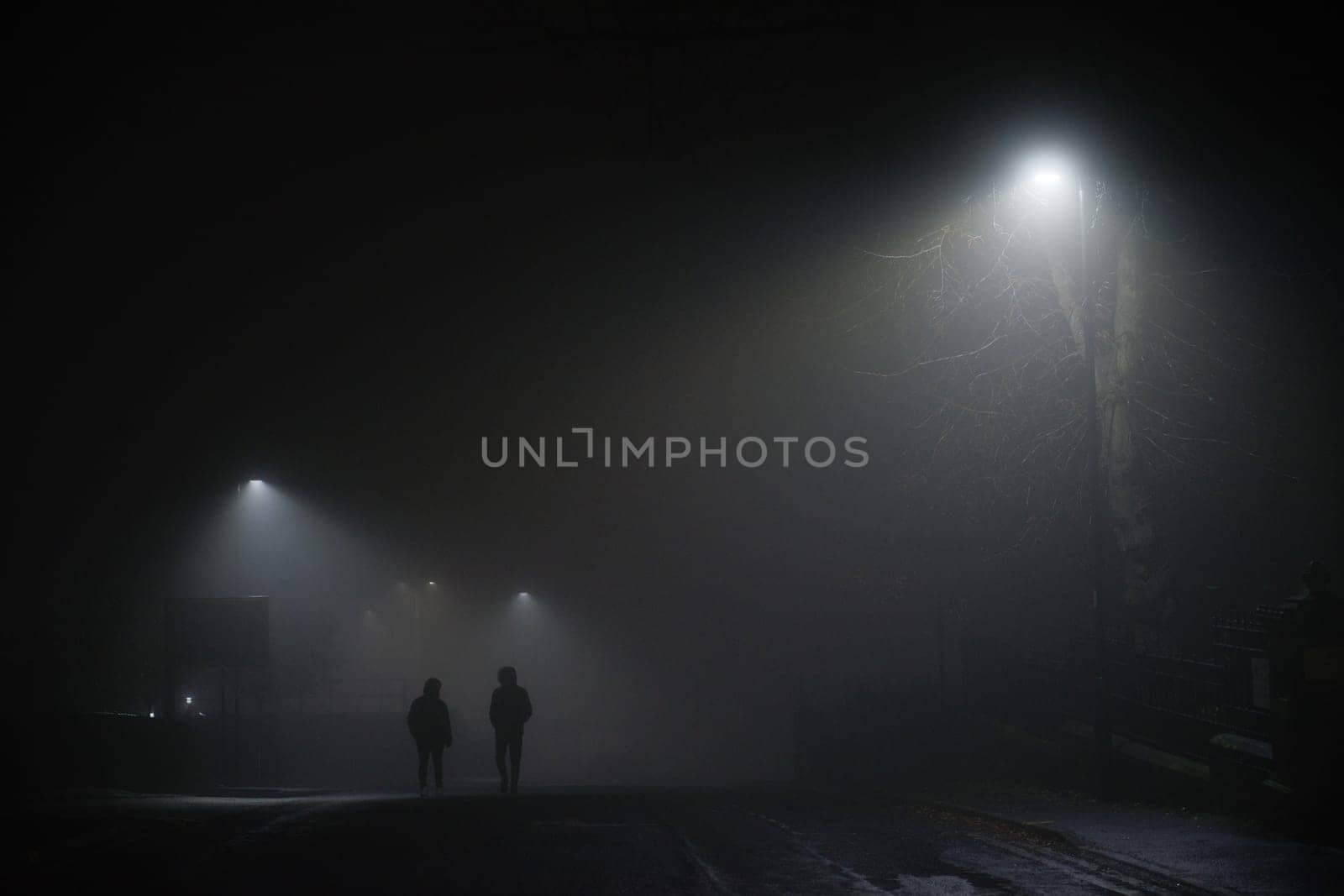 Urban winters scene of two men walking under street lights in extremely foggy conditions. Moody or spooky feel, both wear black coats with hoods up.
