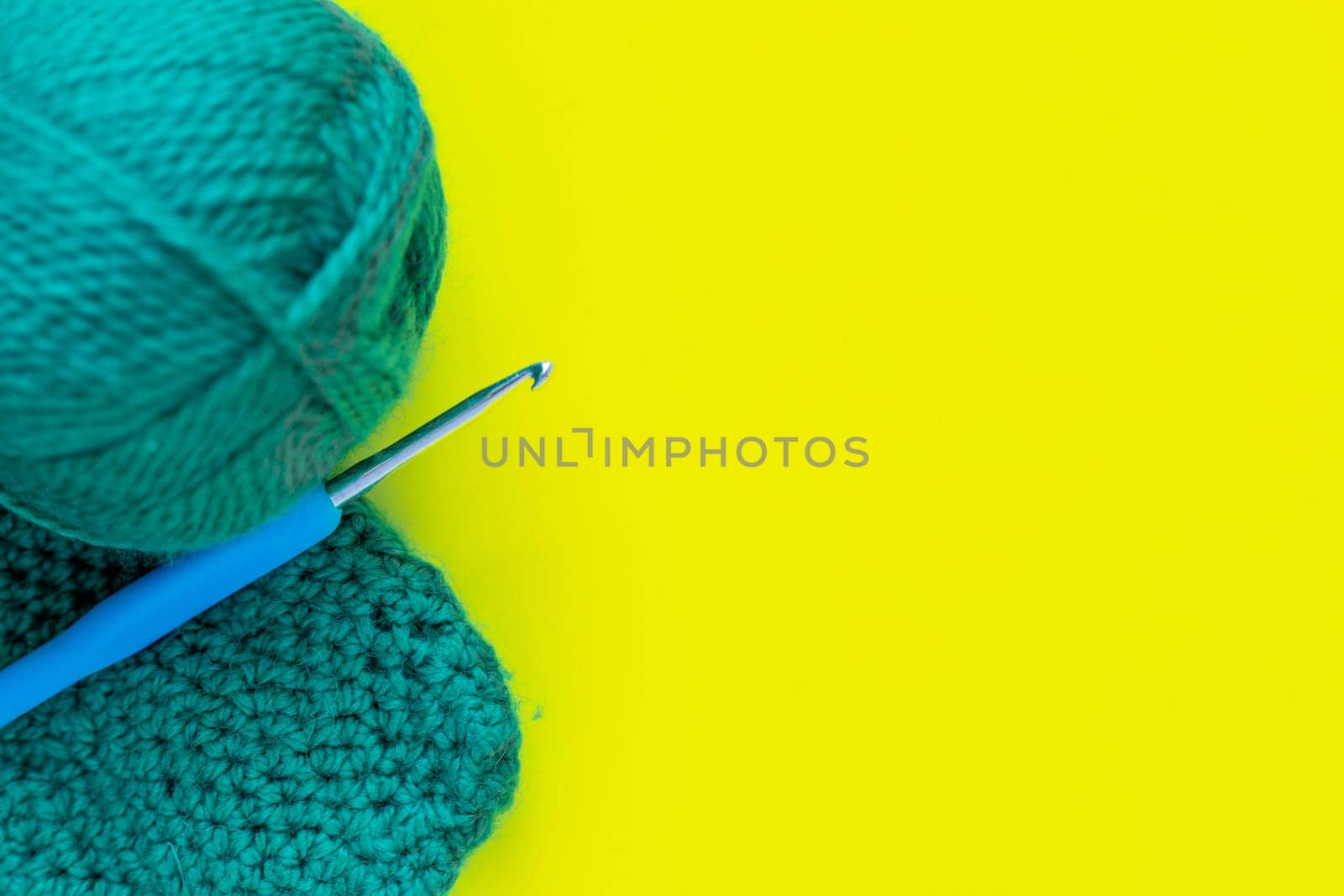 A metal hook with a blue handle, a skein of green yarn and a part of a knitted fragment lies on the side of the photo on a yellow background.