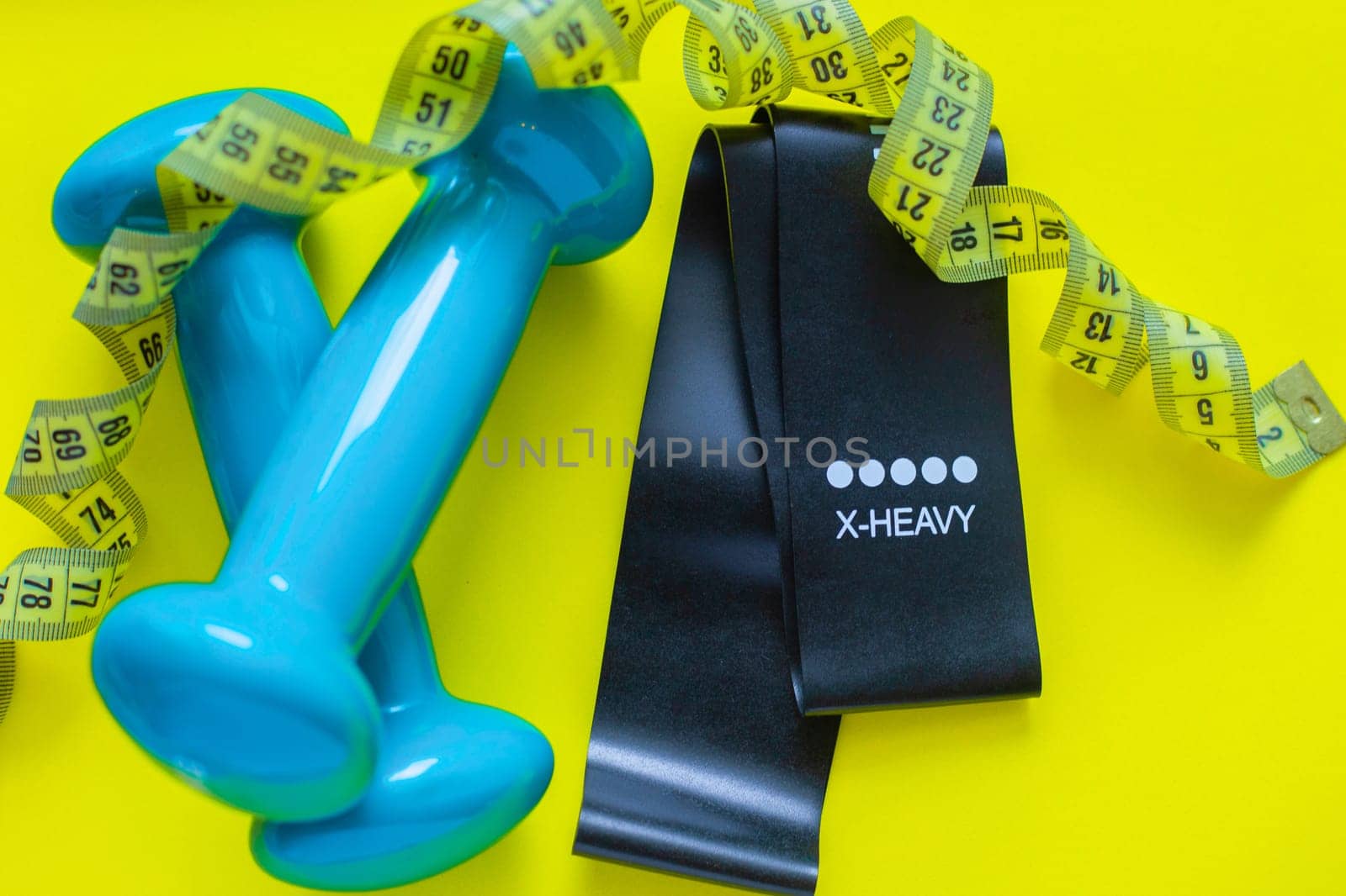 A black fitness elastic band and two blue dumbbells lie on a yellow background, a measuring tape lies above them in rings, close-up. flat lay, copy space, isolate. High quality photo