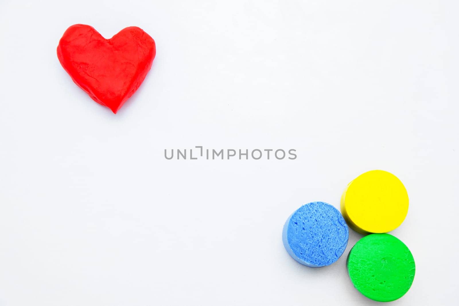 A heart made of red dough for modeling lies on top of the photo, and yellow, green and blue circles lie diagonally on a white background. High quality photo