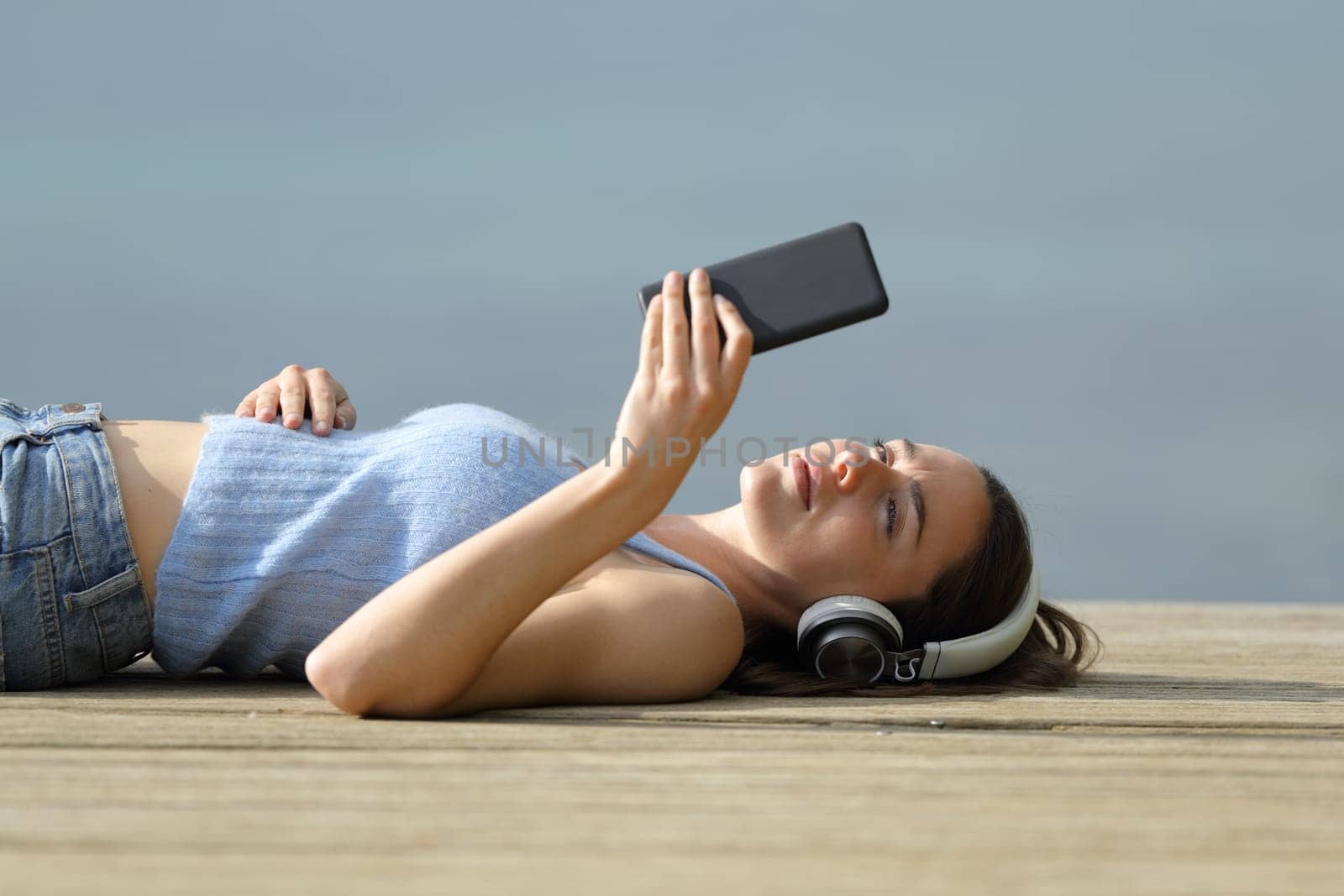 Serious woman lying on pier listening to music with smart phone