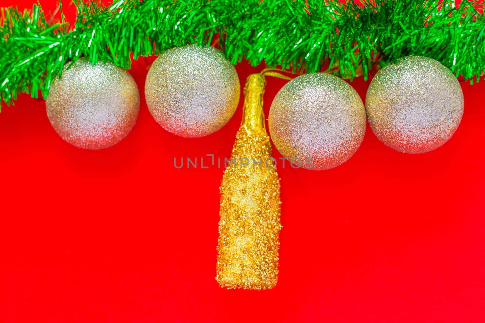Christmas balls of silver and a golden bottle of champagne hang on a green fluffy garland of tinsel green on top of the photo on a red background. top view, flat lay, copy space, isolate. High quality photo