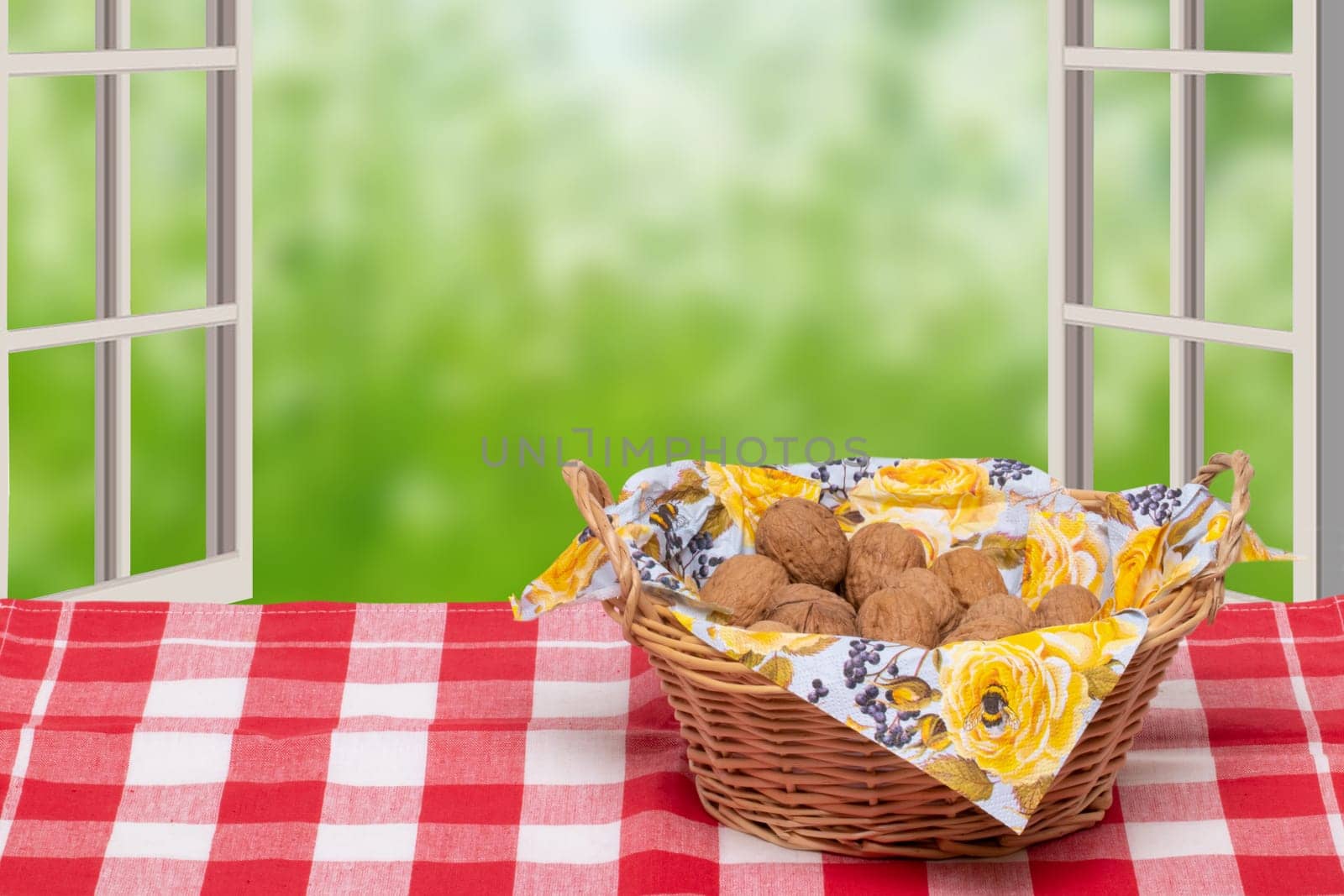 A wicker basket with a napkin full of walnuts on a table with a red checkered tablecloth in front of open windows with an abstract blurred natural green background. Macro.