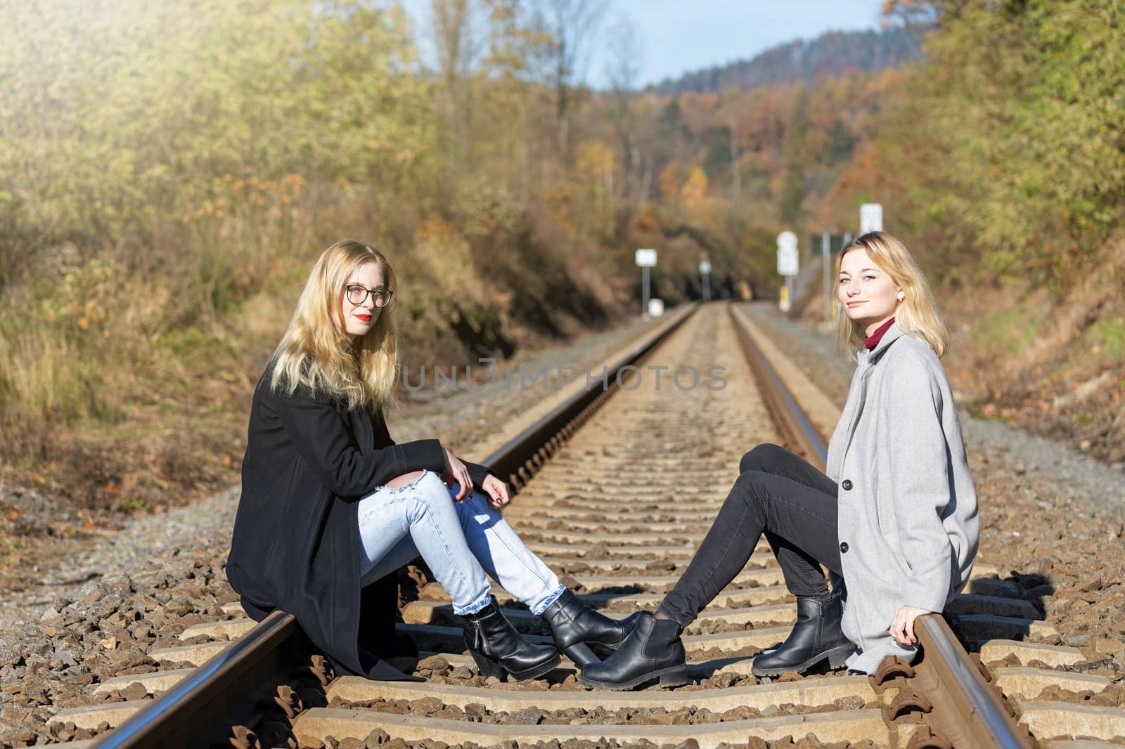 Two attractive young girls are sitting on rail in autumn nature. Horizontally.