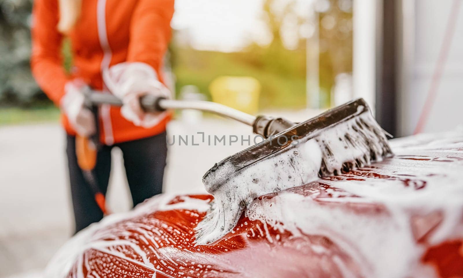 Young woman cleaning her car in self serve carwash. Closeup detail on white soap foam and brush moving over roof.