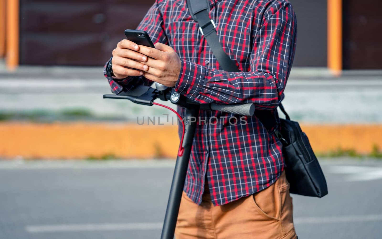 Young man wearing shirt leaning on electric scooter handlebar, holding mobile smartphone in his hands, closeup detail.