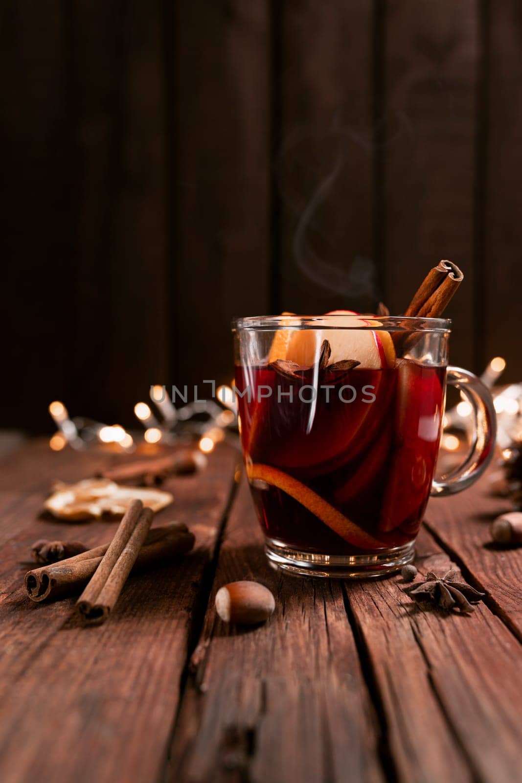 Christmas time concept photo. Glass cup or mug with Mulled Wine inside on wooden table and background. Garlands, decorations, cinnamon, hazelnut, dried or sublimated orange, apple, various spices