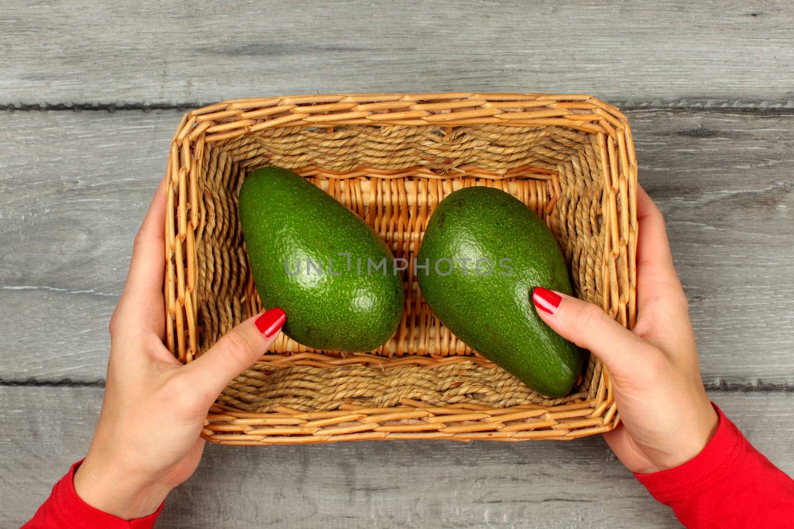 Table top view - young woman hands, red clothing and nails, holding square basket with two avocado, on gray wood desk.