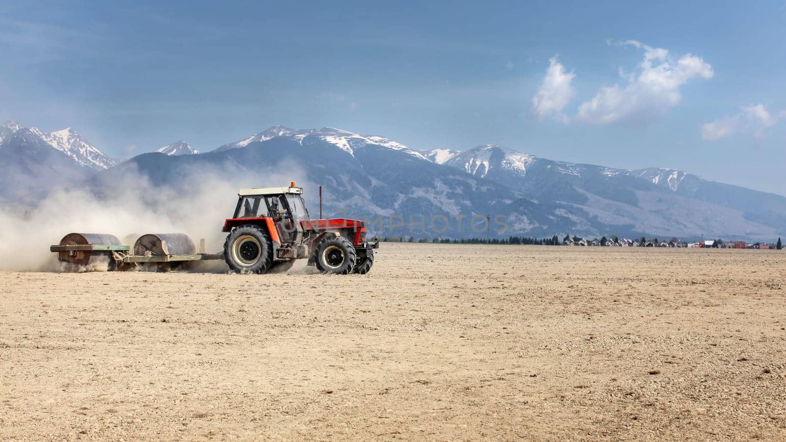Tractor pulling heavy metal roller over dry field on a nice spring day with mountains in background. Soil preparation.