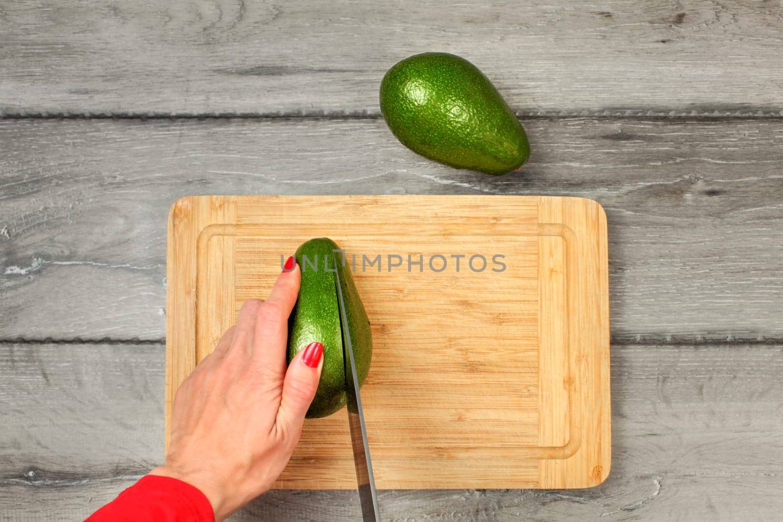 Table top view - young woman hands with red nails holding knife, cutting avocado on chopping board.