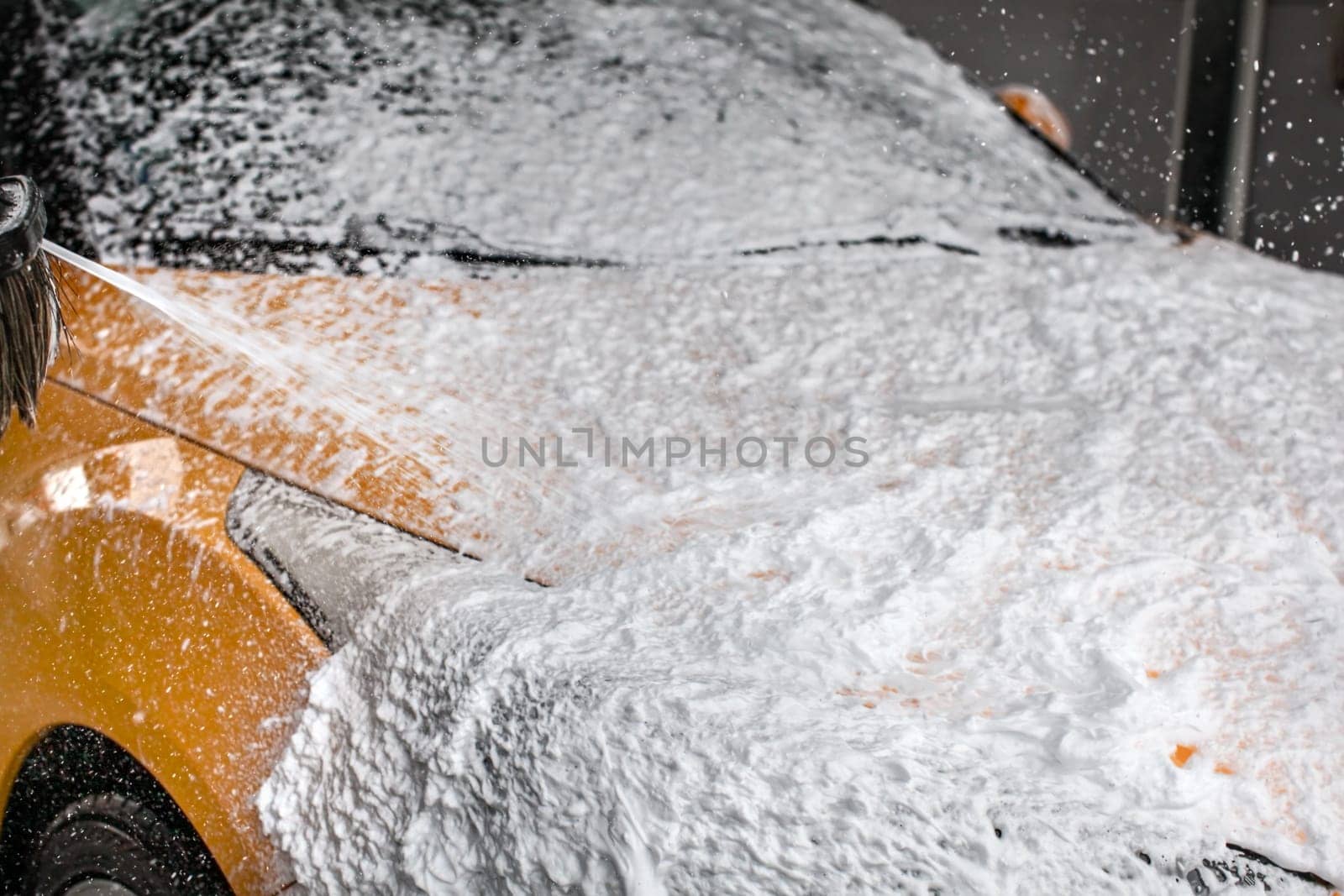 Dark yellow car being washed in carwash. Front completely covered in thick shampoo foam, with more spraying from the brush.