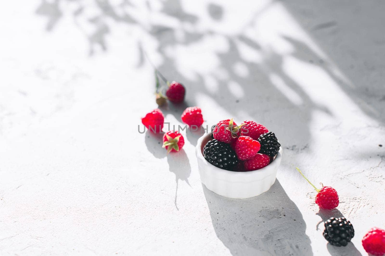 White bowl full with raspberries, blackberry on gray concrete table with leaves, tree branches shadow. Healthy eating concept. Eco, bio farming. Fresh tasty berries on grey background. Quality photo