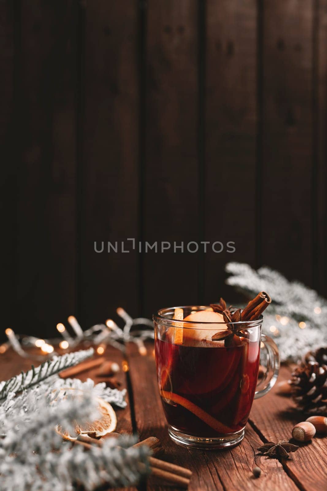 Christmas time concept photo. Glass cup or mug with Mulled Wine inside on wooden table background. Decorations, cinnamon, dried orange, apple, various spices, white fir, cone, anise, aniseed, lights