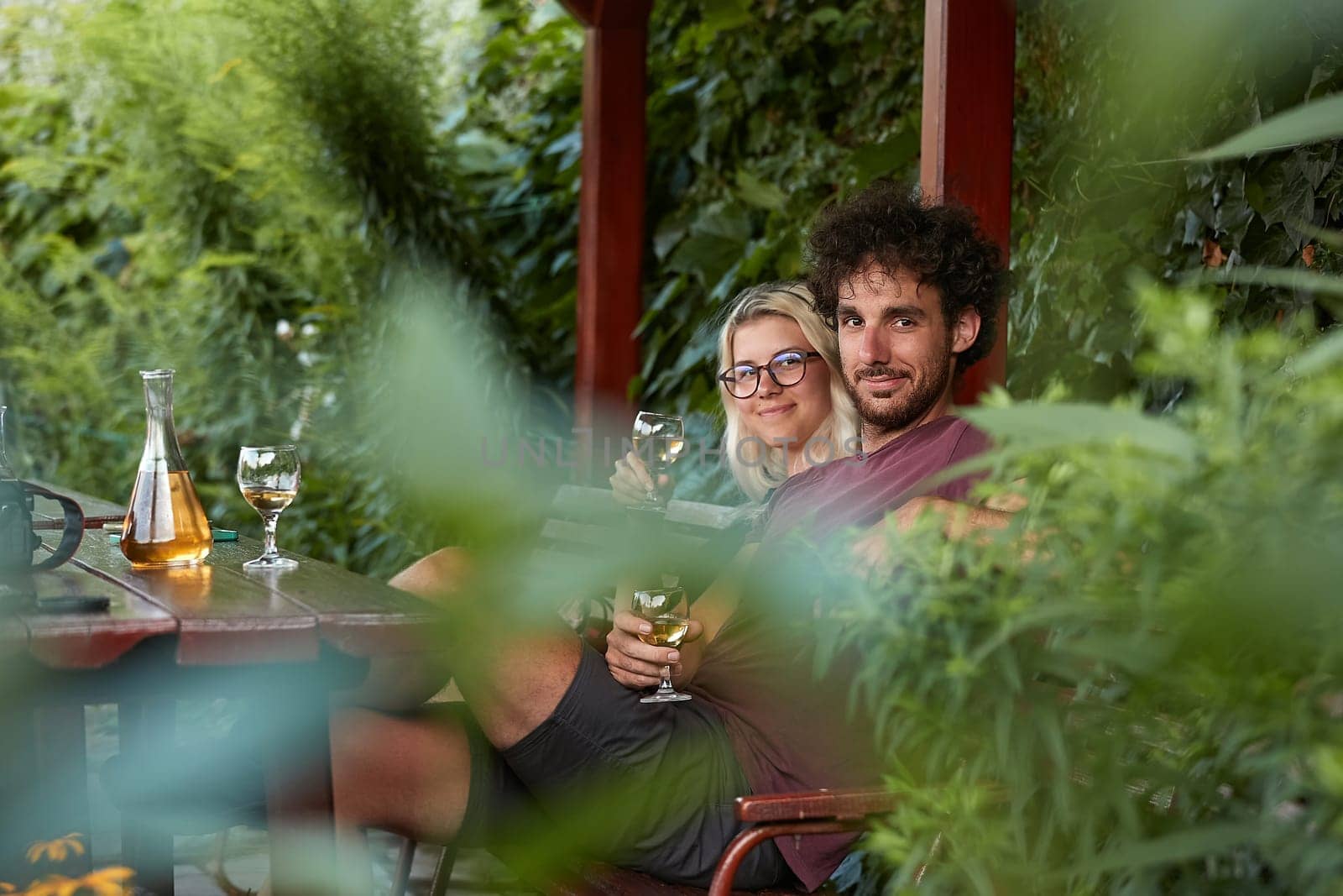 Drinking wine, couple sitting on an outdoor terrace, shot through brances of a green bush in the forground