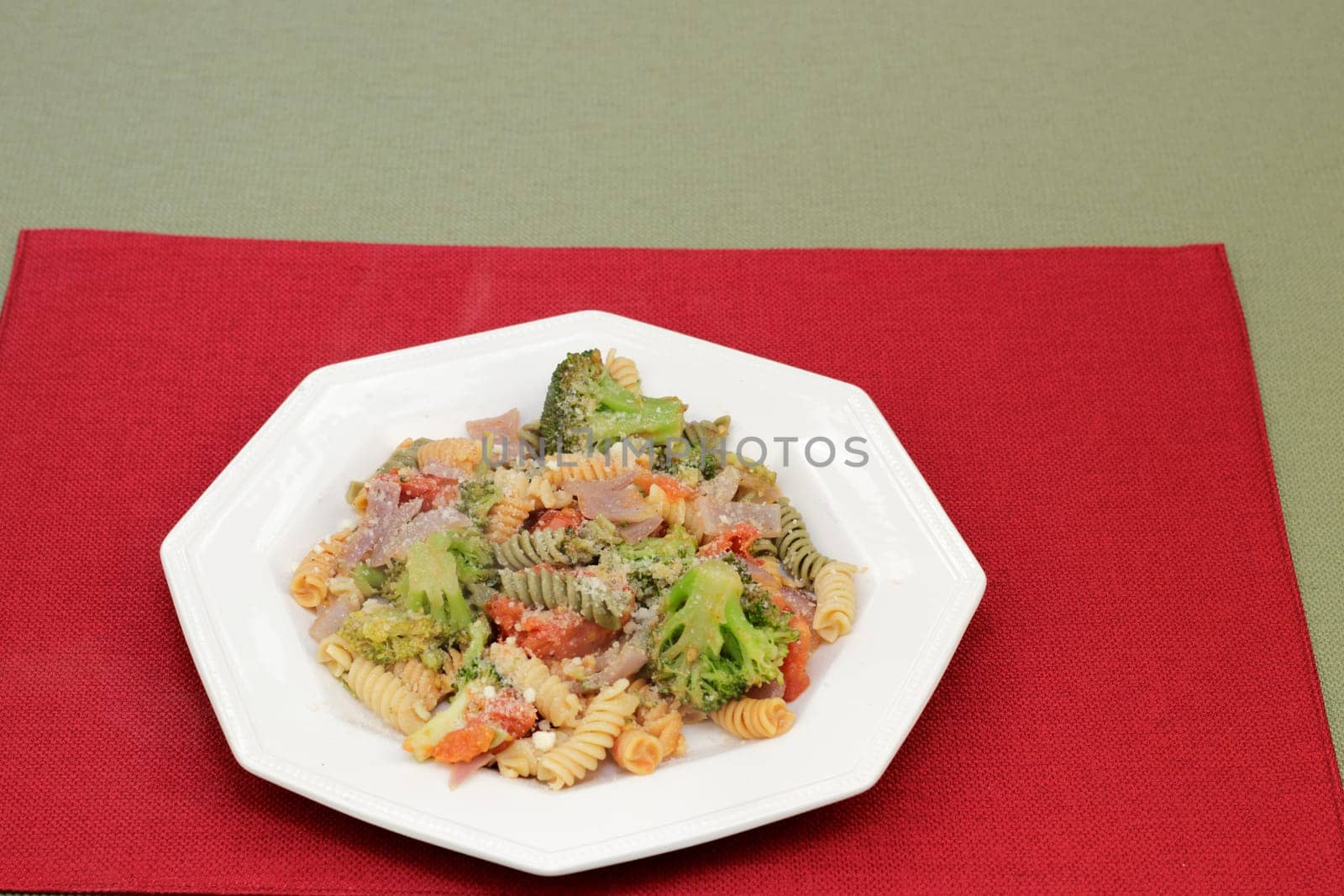 Parmesan cheese topped tri-color vegetable rotini pasta with broccoli, tomato and onions on a plate. Cooked vegetable rotini pasta with mixed vegetables of broccoli, tomatoes and onions with cheese.