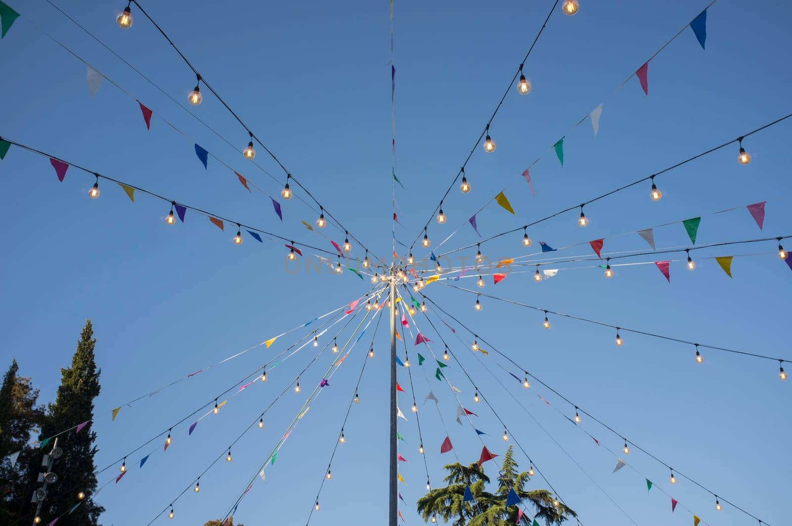 Garland with colorful pennants and light bulbs on pole over blue sky. Outdoors party background