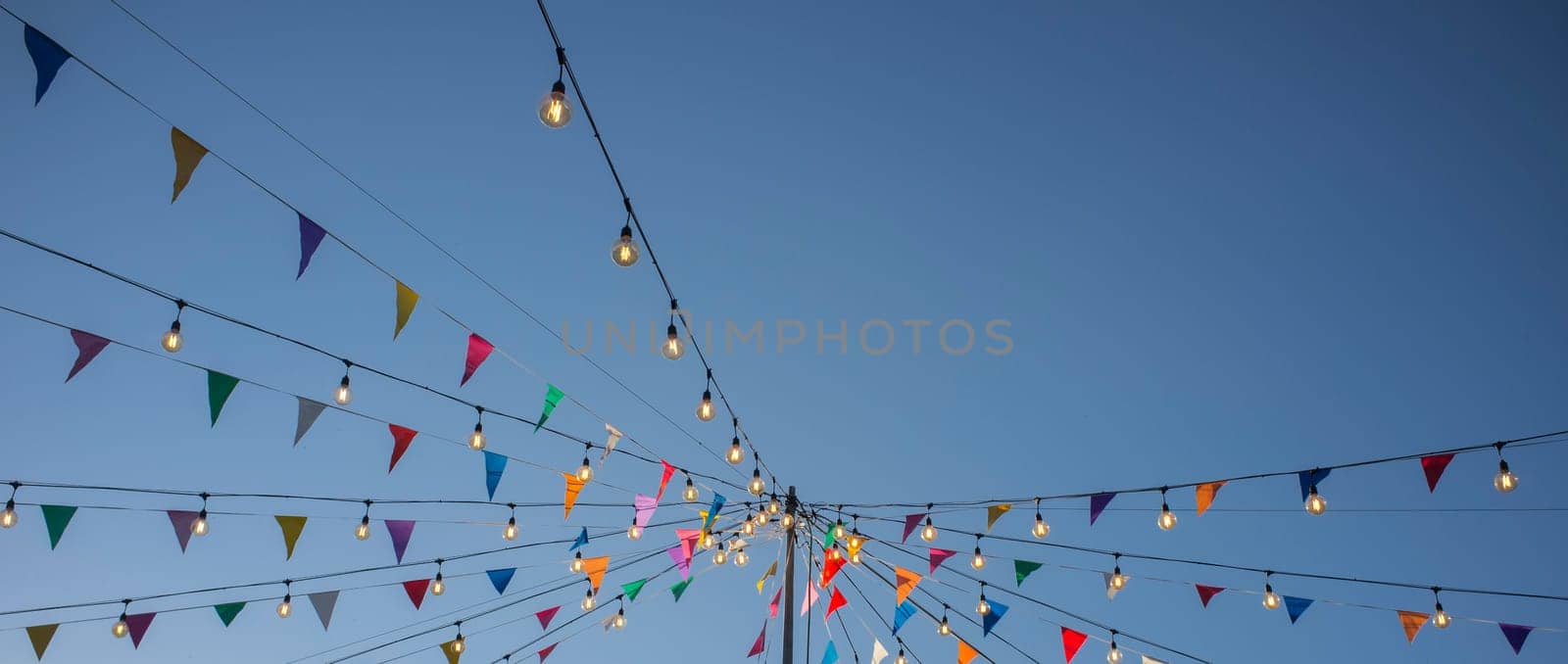Garland with colorful pennants and light bulbs on pole over blue sky. Long format