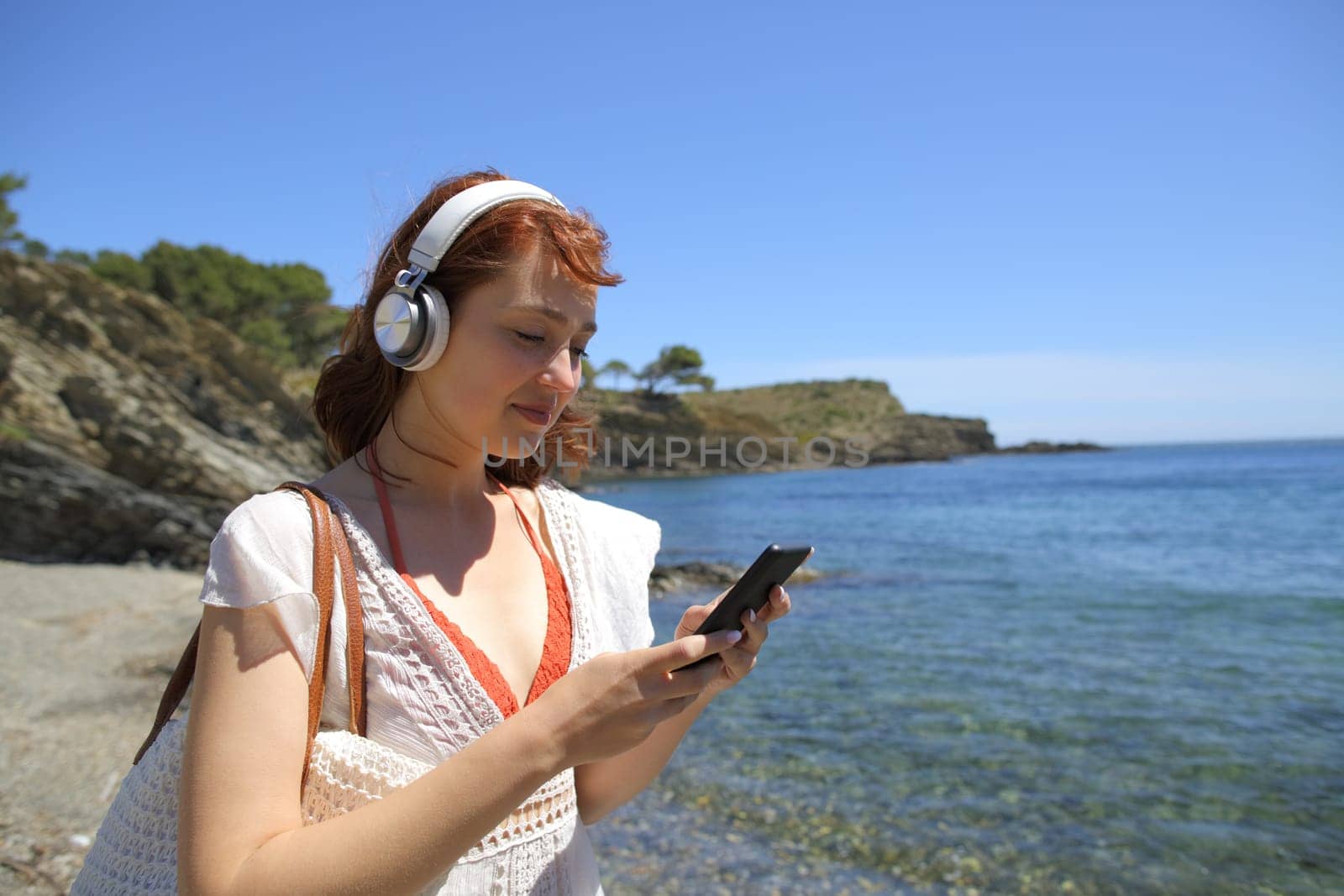 Happy woman wearing wireless headphones using smart phone listening to music on the beach