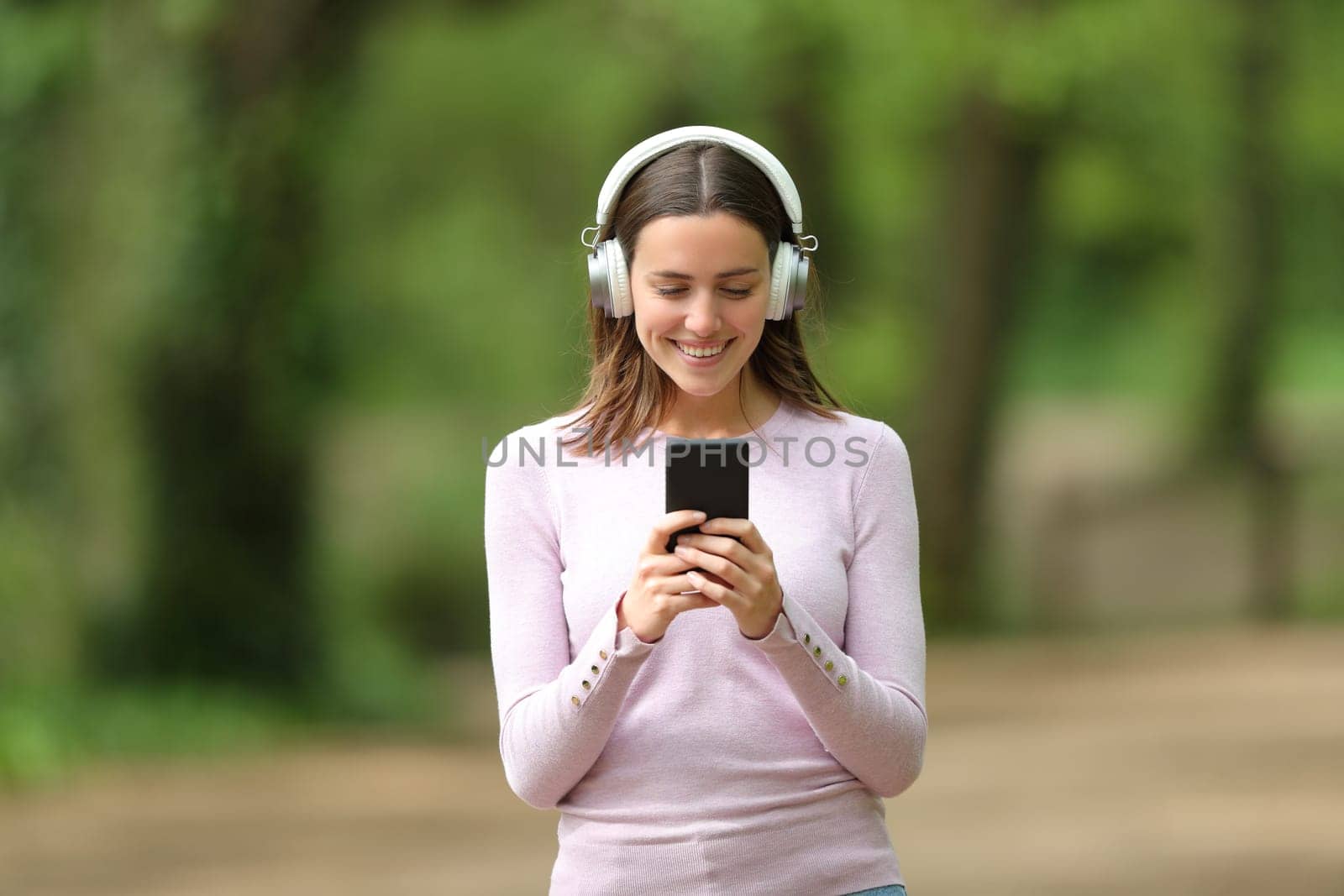 Front view portrait of a happy woman wearing wireless headphones walking listening to music on phone in a park