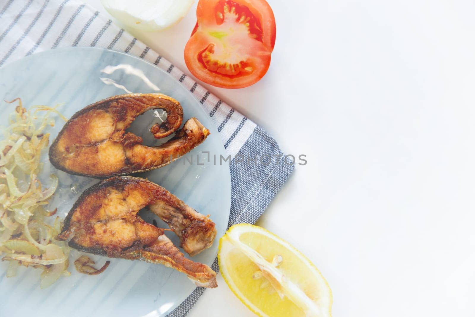 Fried fish with onions lies on a plate, on a waffle towel, next to a lemon and a tomato on a white background. High quality photo
