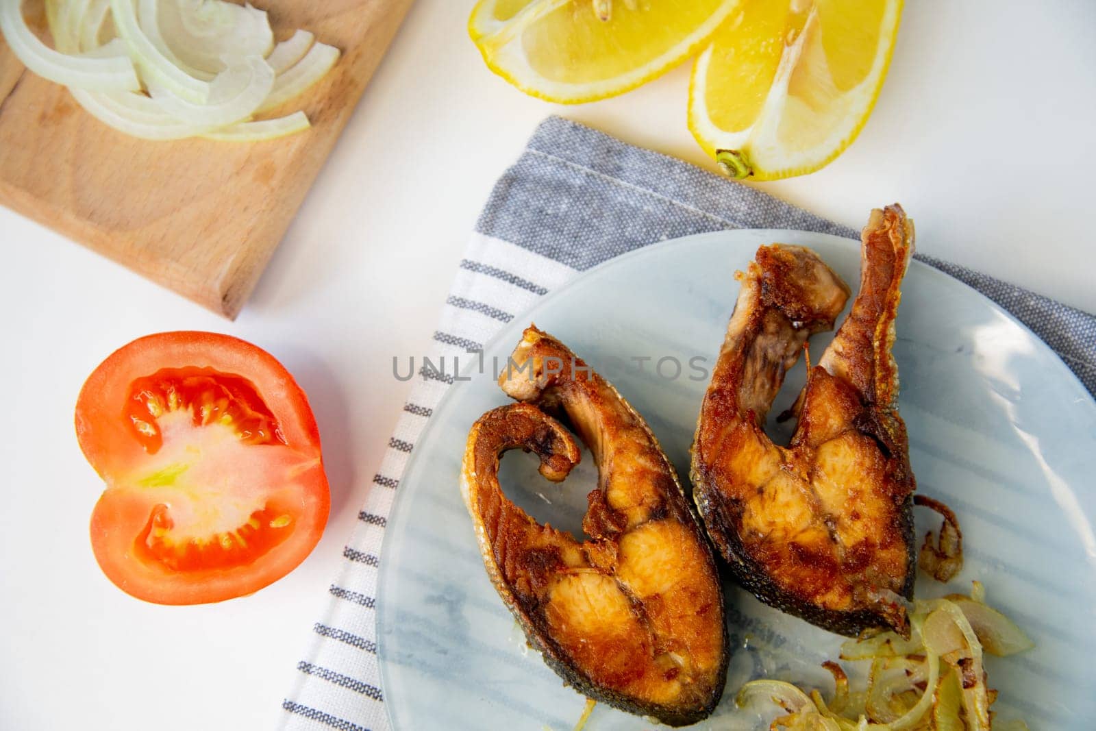 Fried fish with onions is on a plate, on a waffle towel, next to it is a cutting board with chopped onions, lemon and tomato on a white background. High quality photo