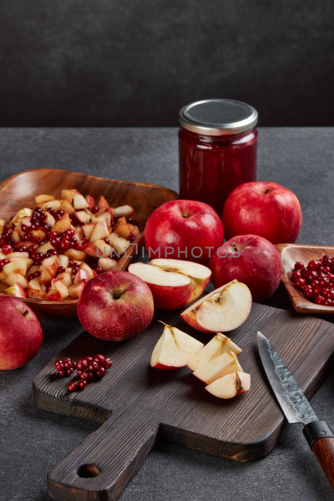 Still life with fresh lingonberries, red appples with knife on black cutting board and jar of homemade jam on dark background. Autumn harvest, Home canning.