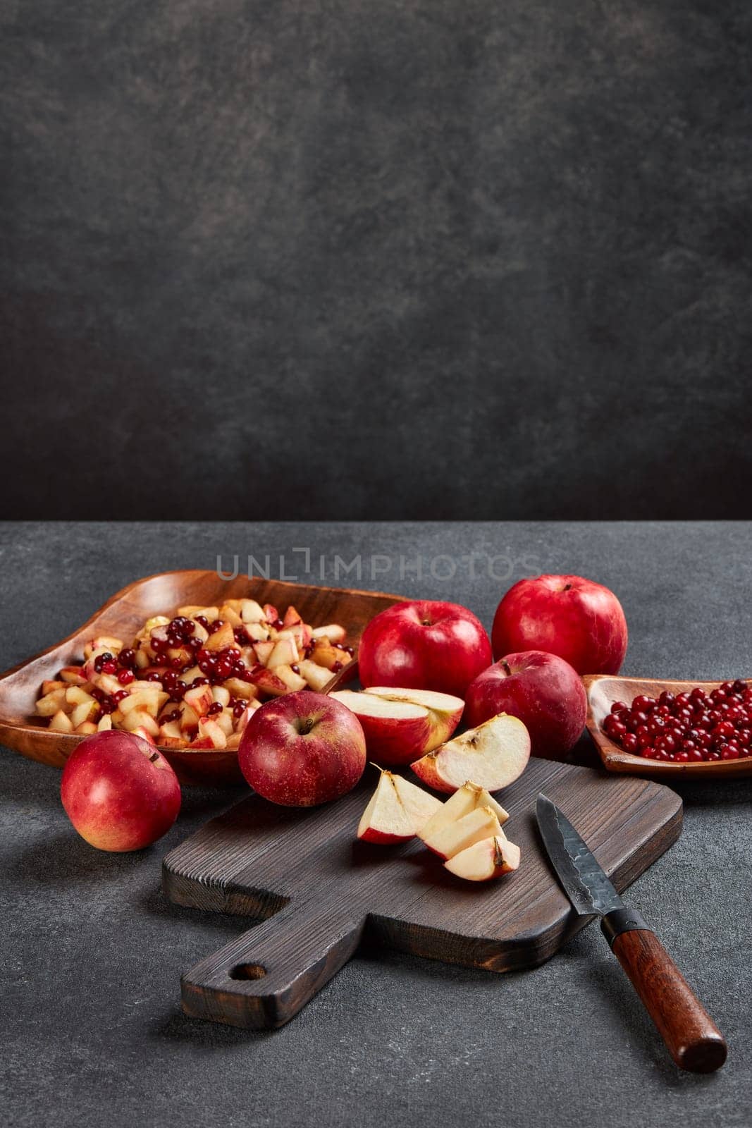 Fruit salad from cowberries and red apples. Still life with apples on black cutting board, ripe lingonberries and fresh fruit salad in wooden bowl on dark background with copy space.