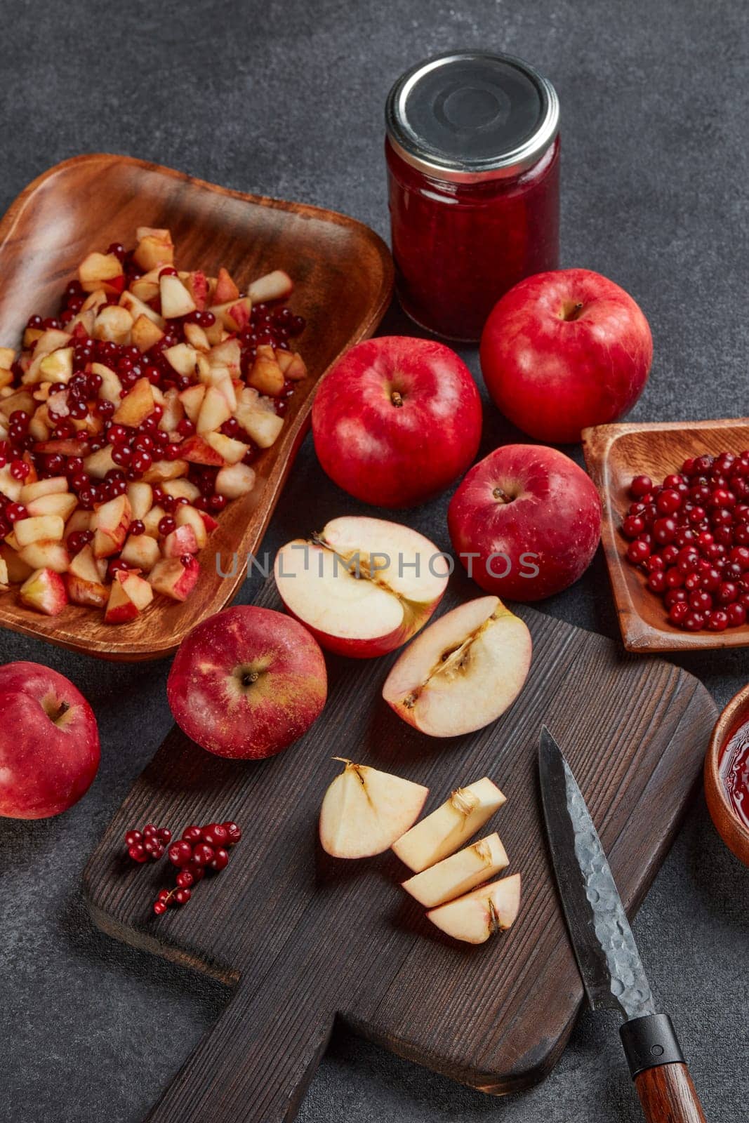 Still life with fresh lingonberries, red appples with knife on black cutting board and  homemade jam on wooden bowl on dark background. Top view. Autumn harvest, Home canning.