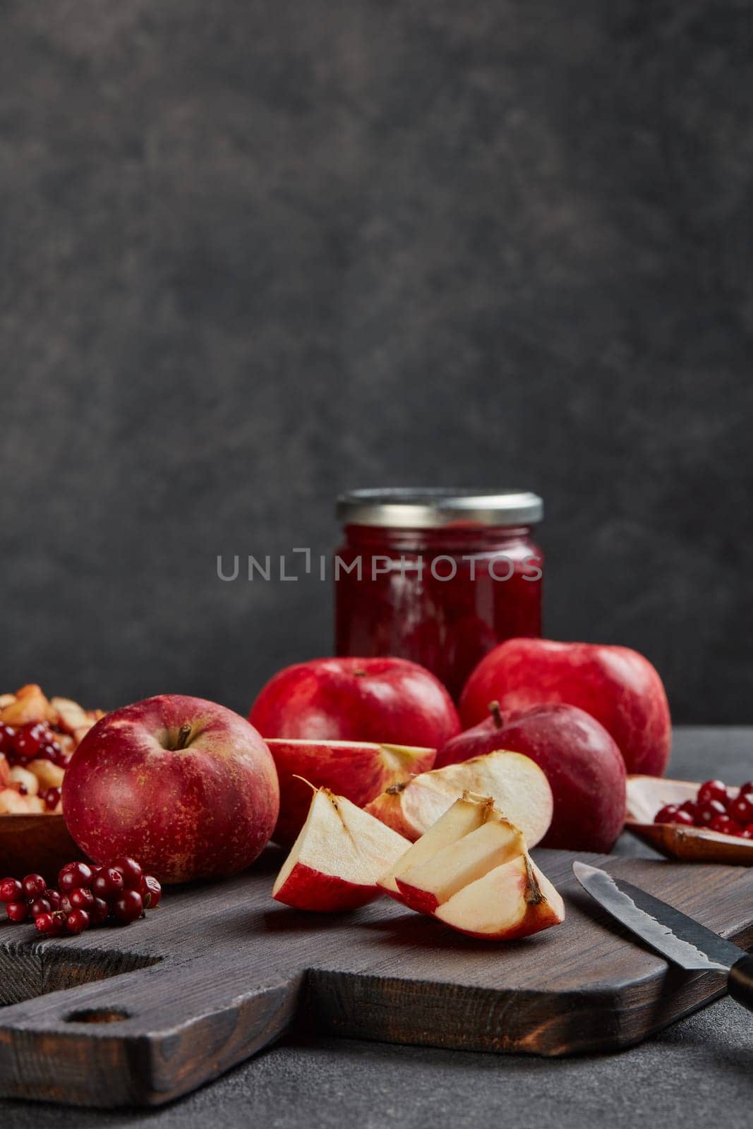 Still life with fresh lingonberries, red appples with knife on black cutting board and jar of homemade jam on dark background with copy space. Autumn harvest, Home canning.