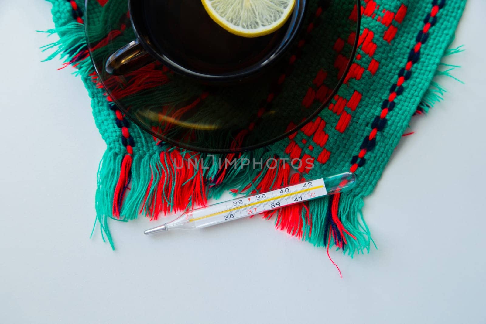 A cup of tea with lemon stands on a dark saucer, which stands on a knitted green-red scarf, next to it lies a thermometer on a white background. . High quality photo