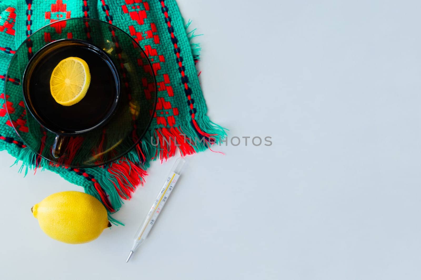 A cup of tea with lemon stands on a dark saucer, which stands on a knitted green-red scarf, next to it lies a thermometer and a lemon on a white background. High quality photo