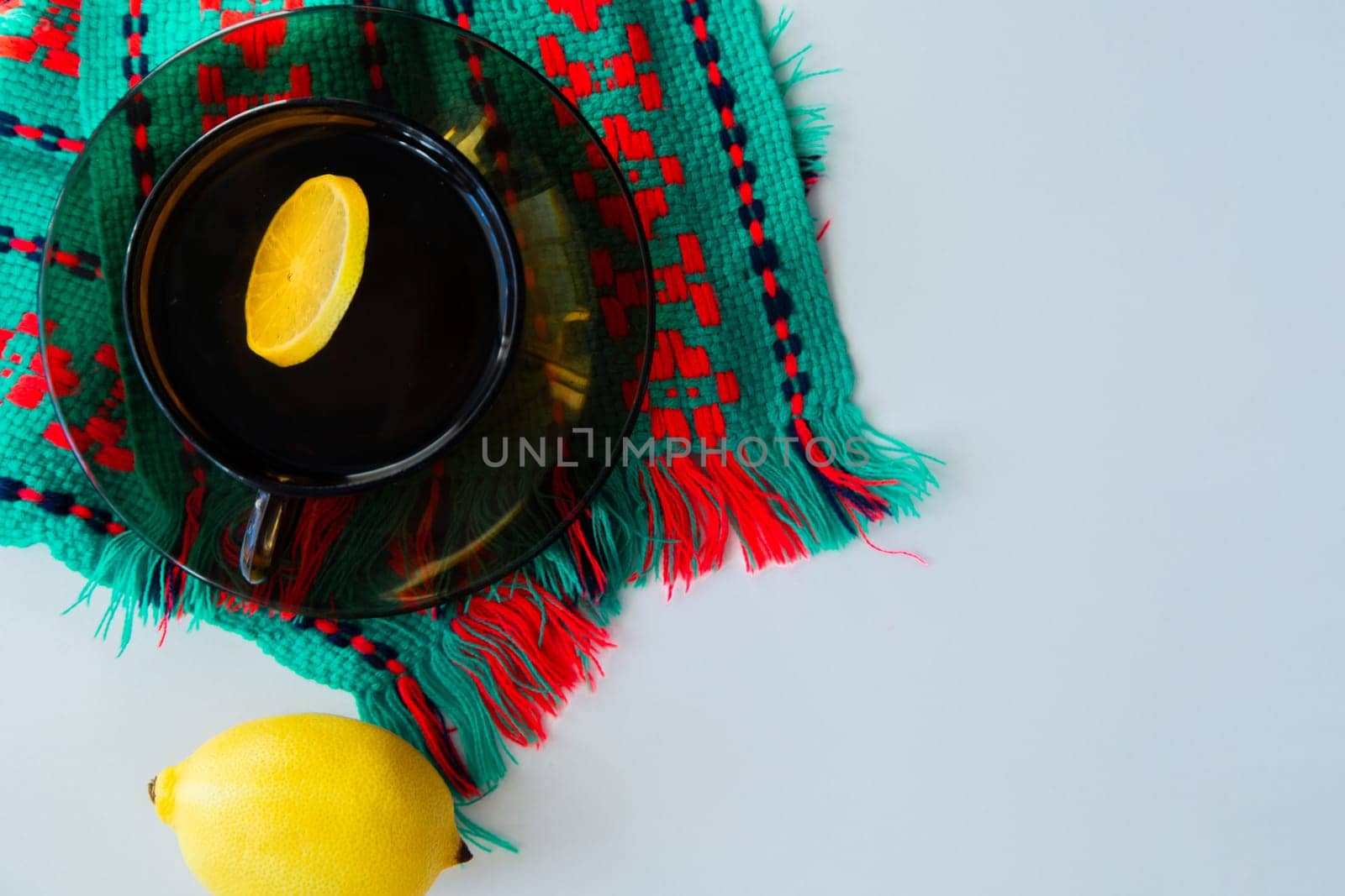 A cup of tea with lemon stands on a dark saucer, which stands on a knitted green-red scarf, next to it lies a thermometer and a lemon on a white background. High quality photo