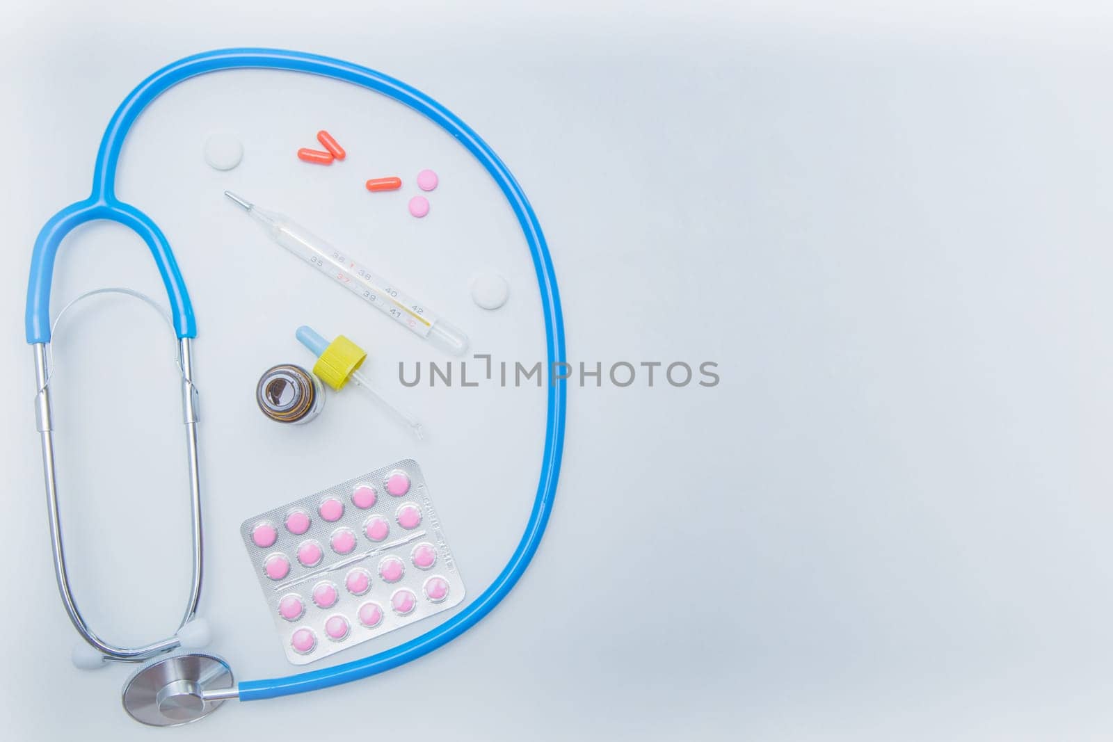 The stethoscope lies on a white background in a circle, inside it is a thermometer, medicines and drops from a runny nose. . High quality photo