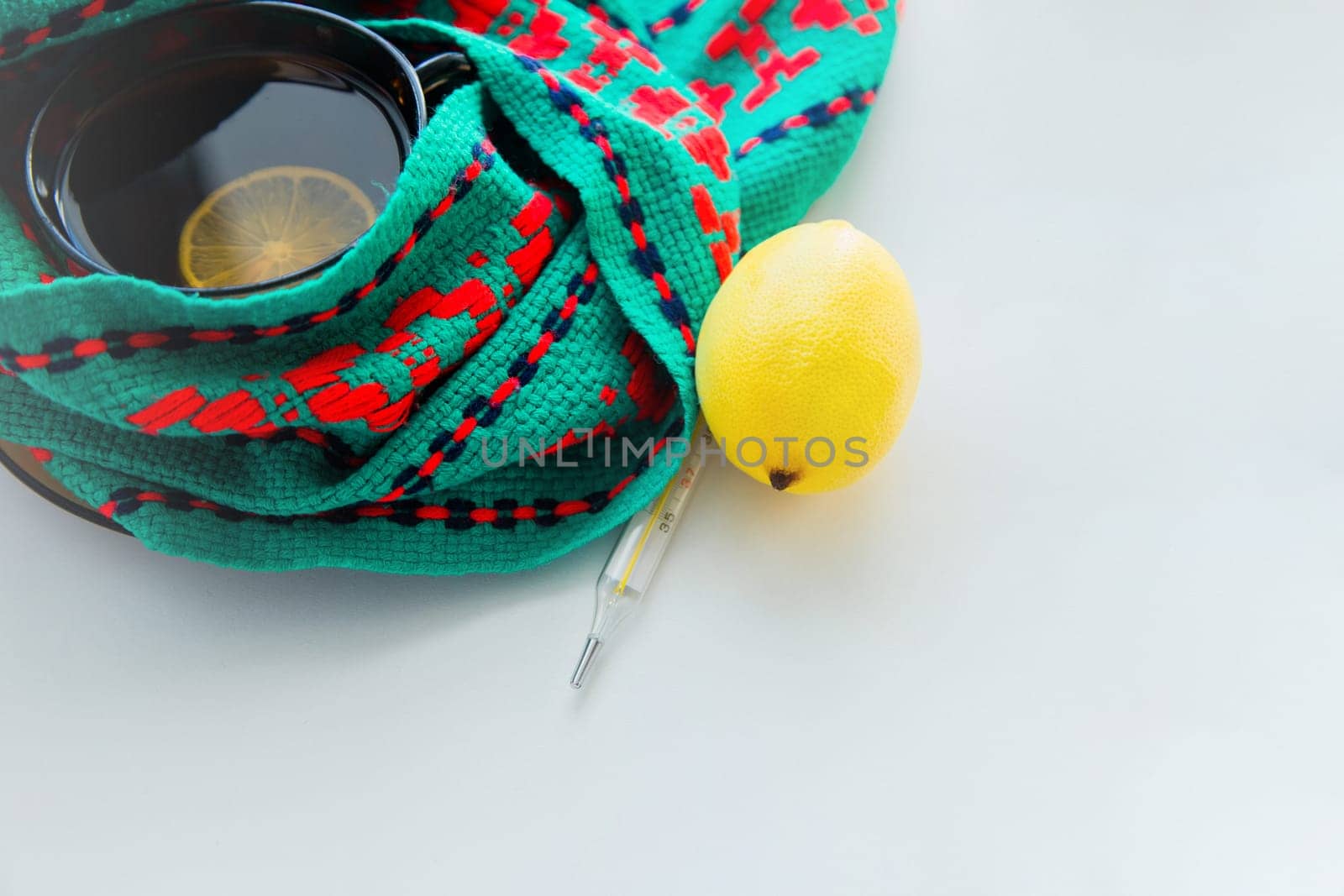 A cup of tea with lemon stands on a knitted green and red scarf, there is a thermometer and a lemon next to it. High quality photo