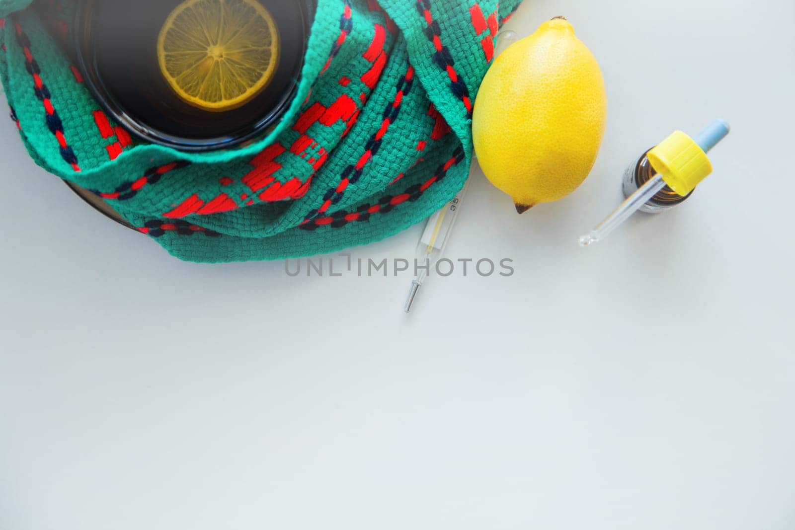 A cup of tea with lemon stands on a knitted green and red scarf, next to it lies a thermometer, lemon and drops from a runny nose. High quality photo