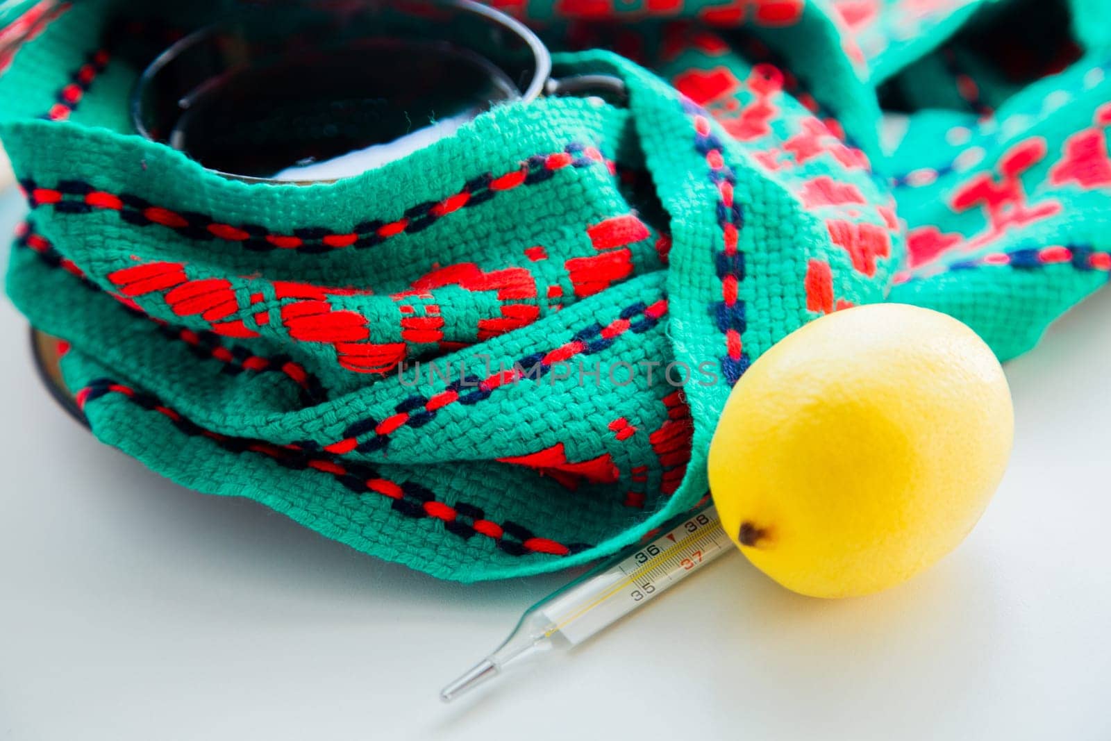 A cup of tea with lemon stands on a knitted green and red scarf, there is a thermometer and a lemon next to it. High quality photo