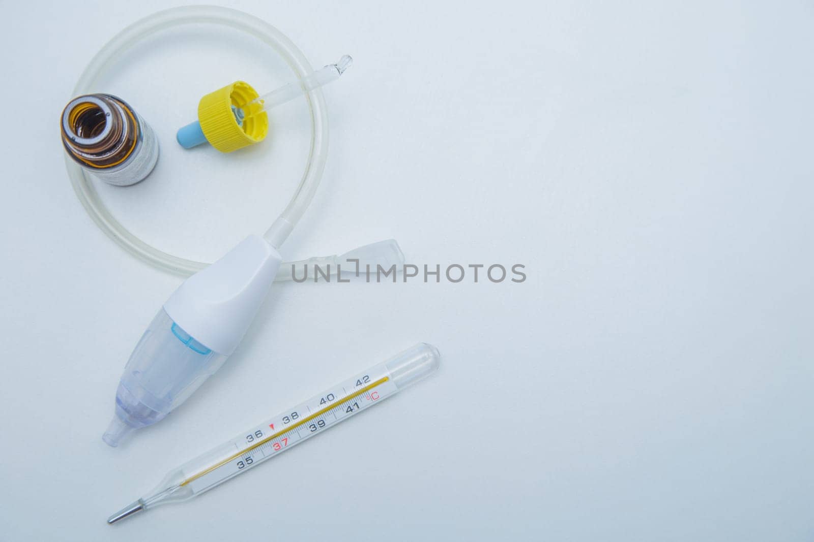 The children's aspirator lies rolled up in a ring on a white background, drops from a runny nose with a pipette in its interior, a thermometer with a place for text lies on the side. . High quality photo