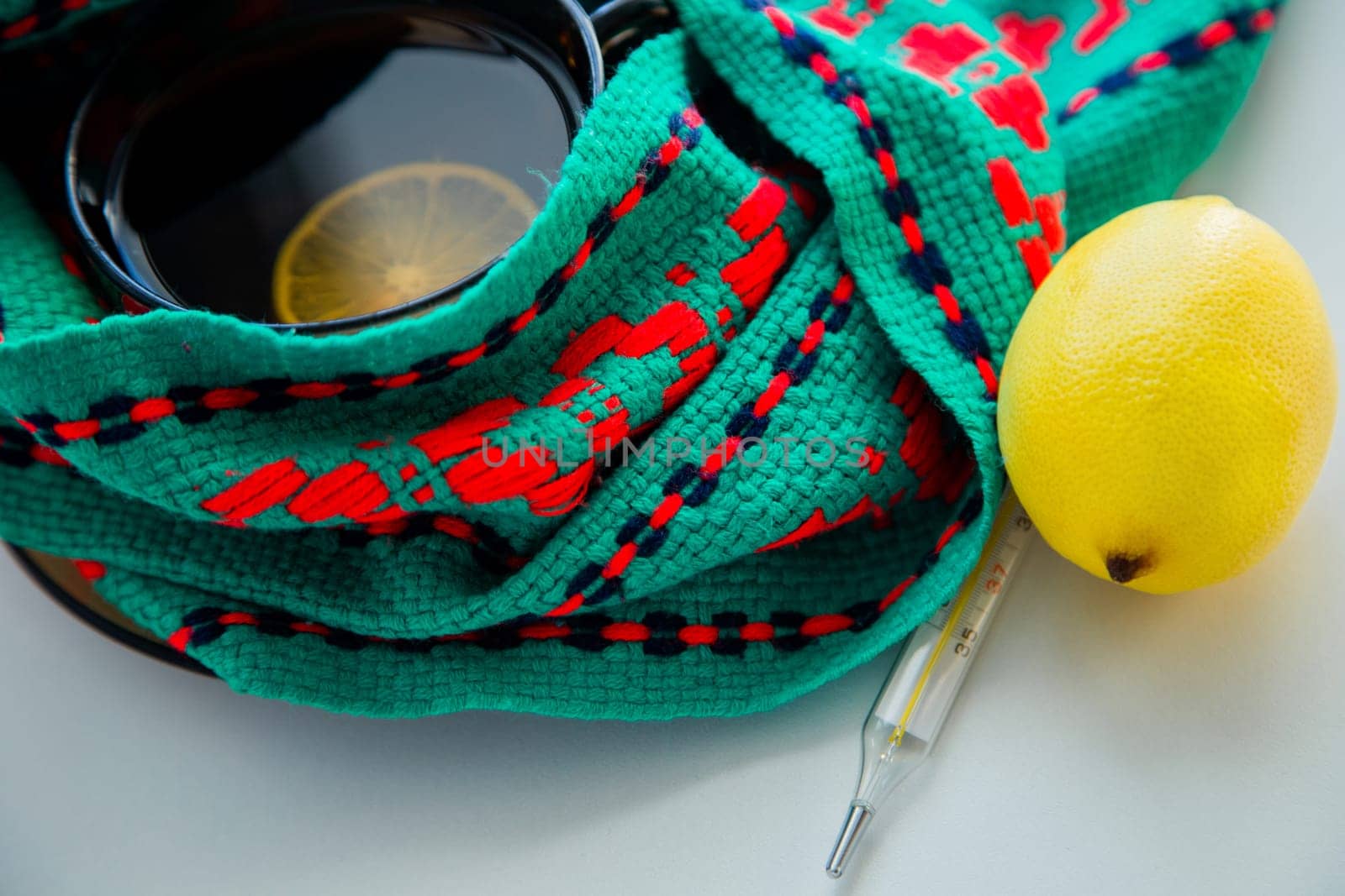 A cup of tea with lemon stands on a knitted green and red scarf, there is a thermometer and a lemon next to it. High quality photo