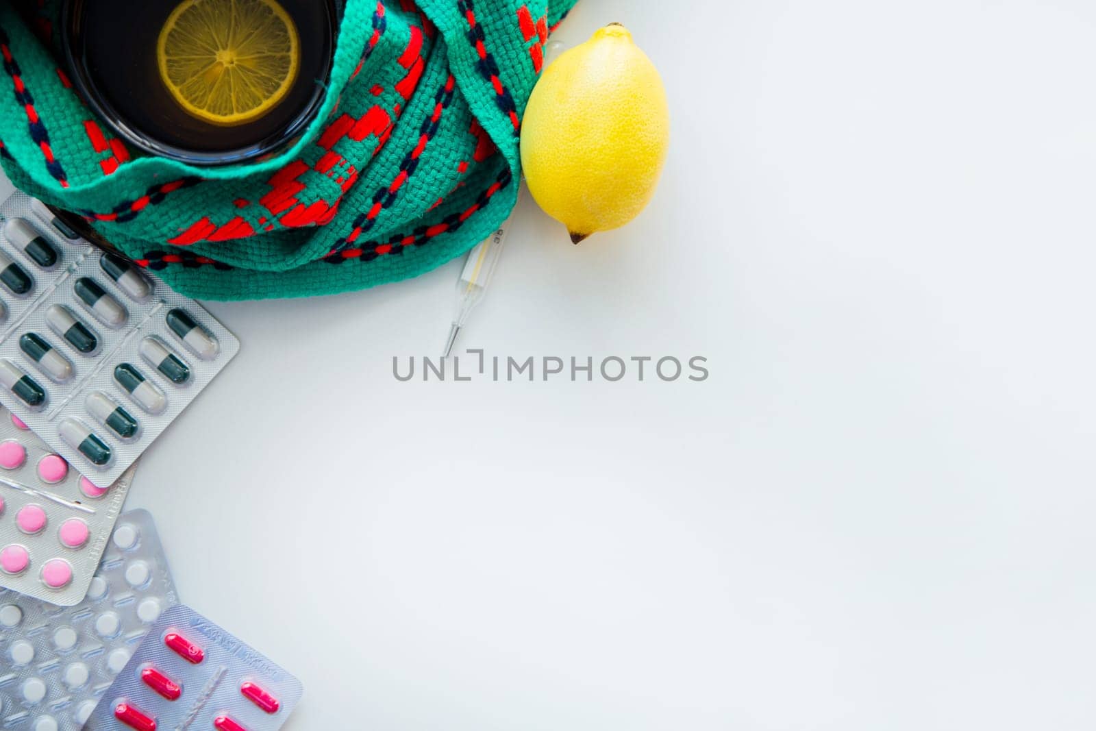 A cup of tea with lemon stands on a knitted green and red scarf, there is a thermometer next to it, a lemon and pills on the side. . High quality photo