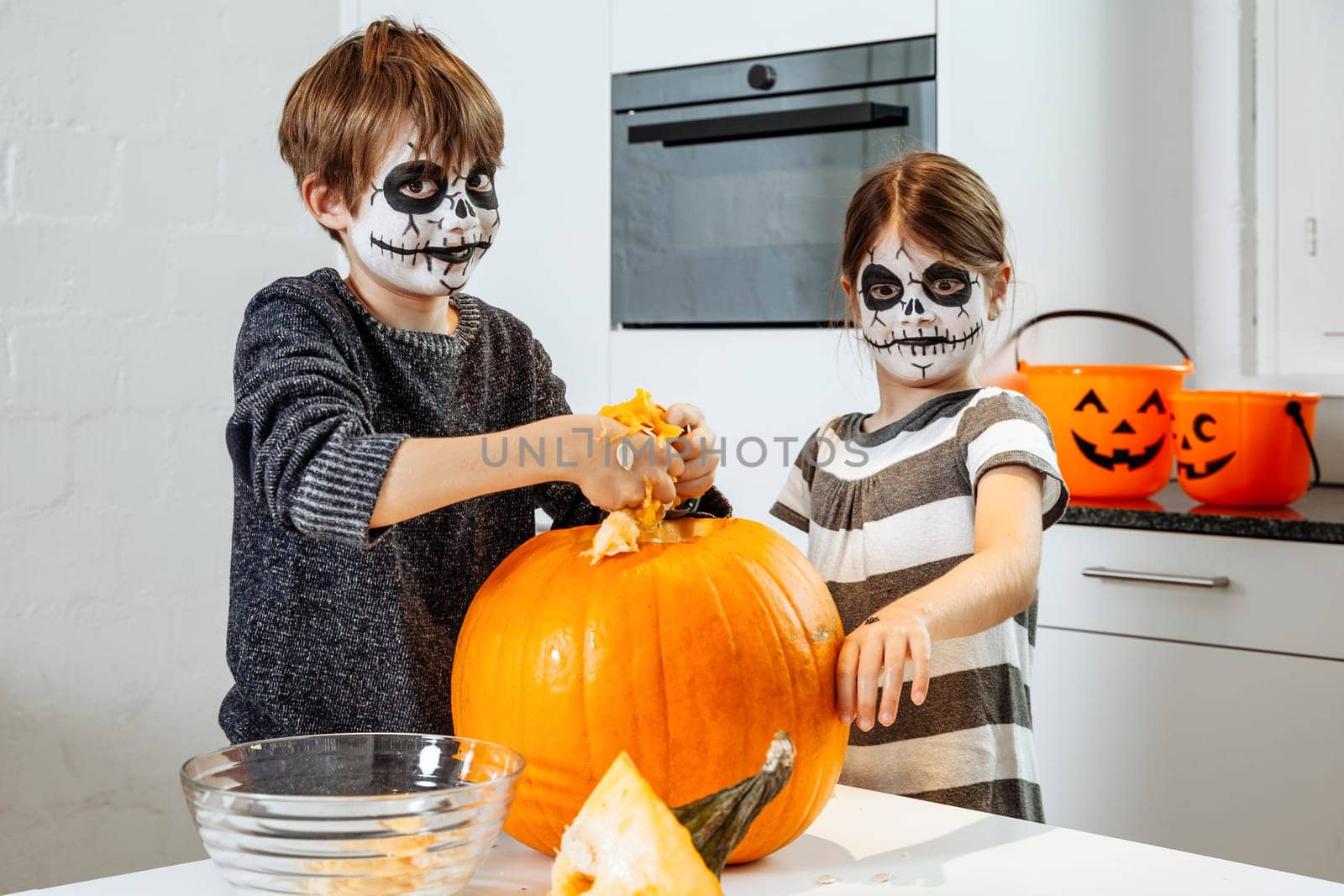 Two young children with skull face paint carving a pumpkin in their kitchen.